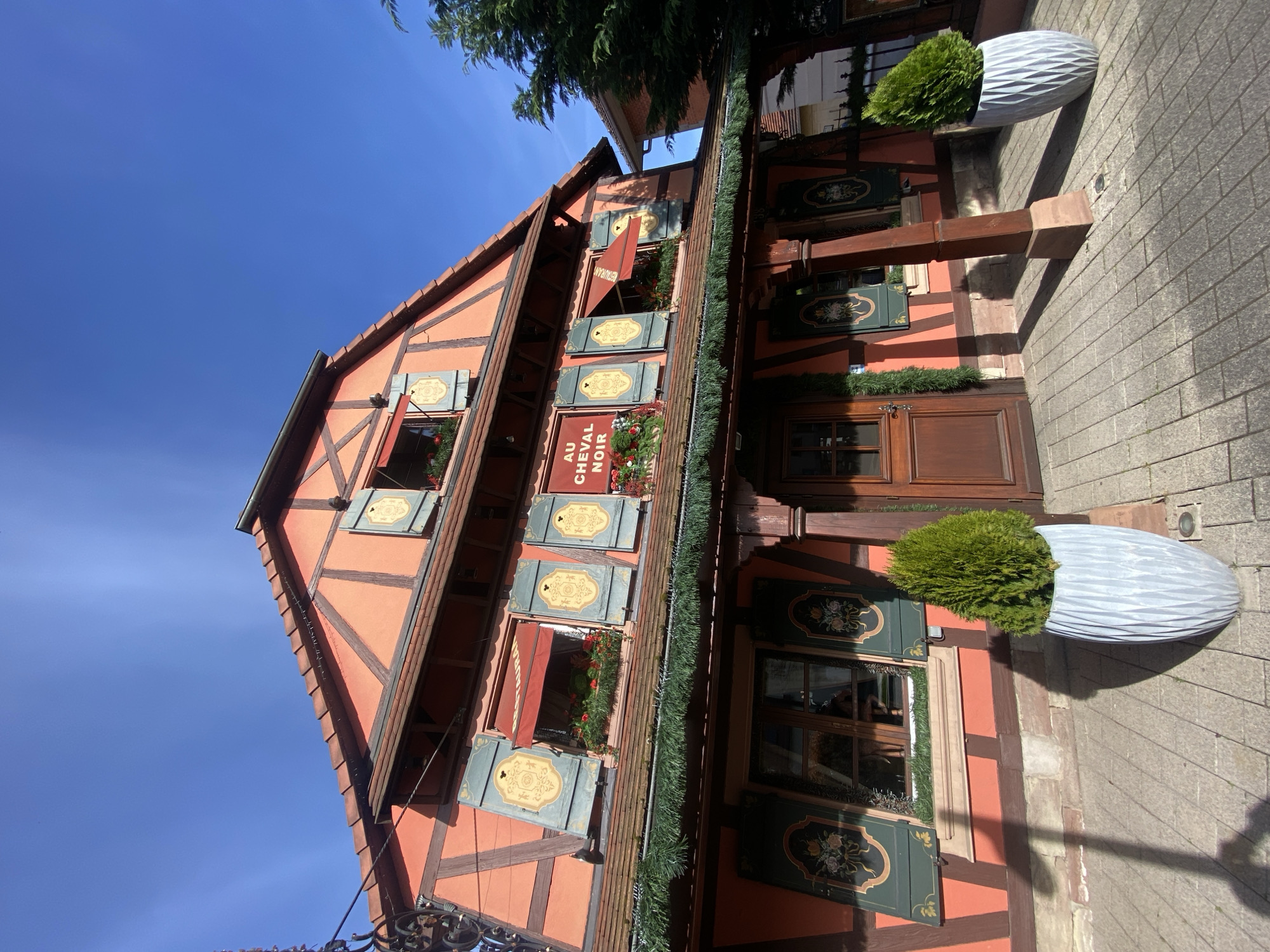 The image depicts a building in Hoenheim, France. The structure is characterized by its traditional half-timbered construction, with a red-orange facade and exposed dark-brown wooden beams. The building features multiple windows with decorative green shutters. A sign reading "Au Cheval Noir" is displayed on the front of the building. Garlands and floral arrangements adorn the windows and facade. Two large, gray-toned planters with green shrubbery flank the building's entrance, which is a wooden door with a small window. The building is set against a clear blue sky. The foreground includes a section of a brick-paved street.