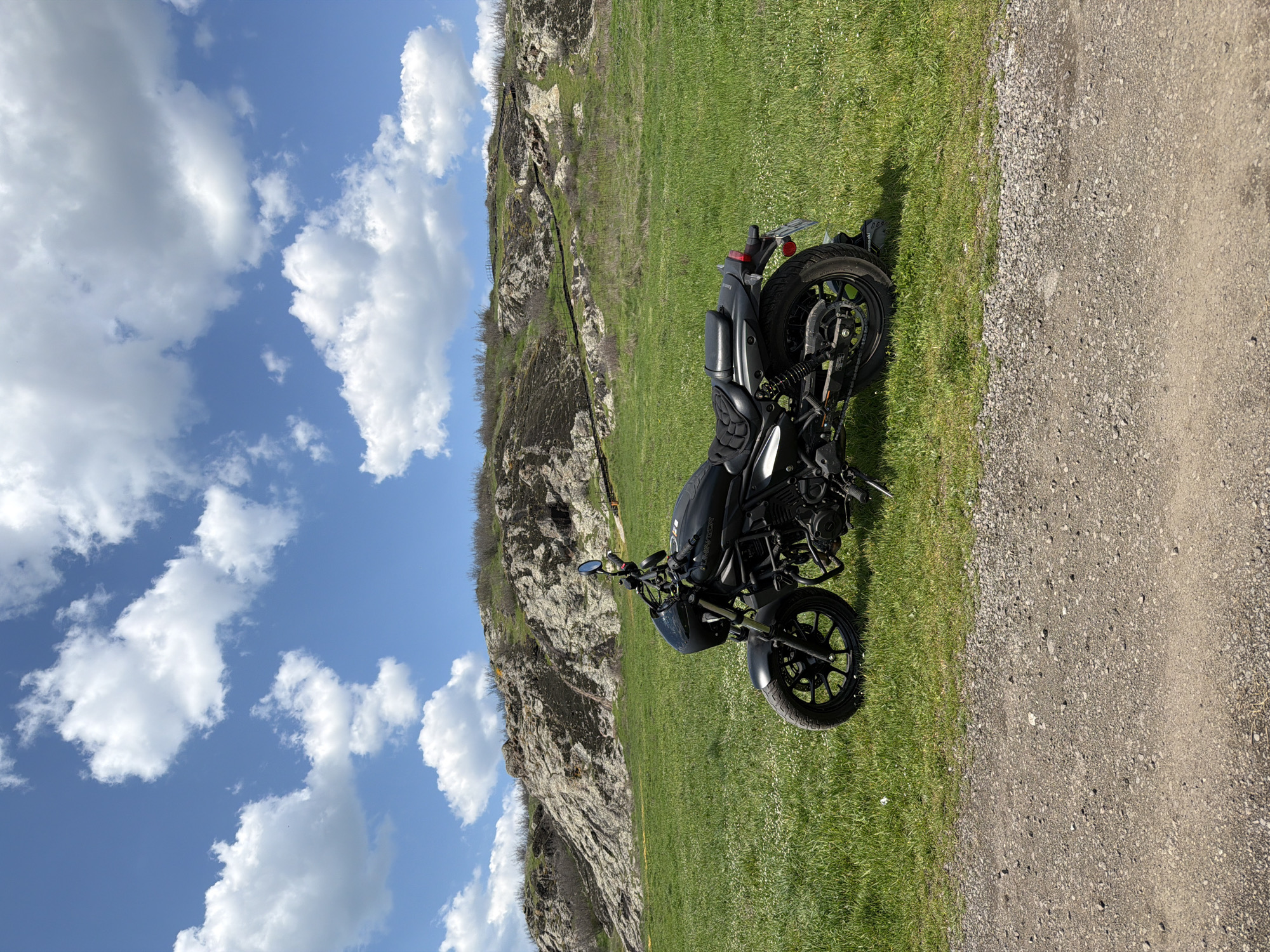 A black motorcycle is parked on a grassy area adjacent to a dirt road. Behind the motorcycle is a rocky hillside. The sky is blue with scattered white clouds. The location is Rusokastro, Bulgaria.