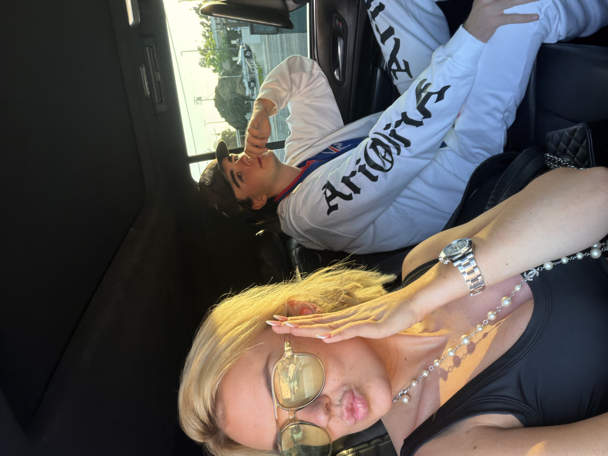 A woman and a man are inside a vehicle, possibly a car. The woman is in the foreground with blonde hair, sunglasses, and a black top. She is wearing a watch on her left wrist, a pearl necklace, and she is posing with her hand near her face and puckered lips. The man is in the background, wearing a baseball cap and a white long-sleeved shirt with black text on the sleeves, and white pants with "ALL" printed on the thigh. The background visible through the car window suggests an urban area with buildings and a street, possibly in Los Angeles, United States.