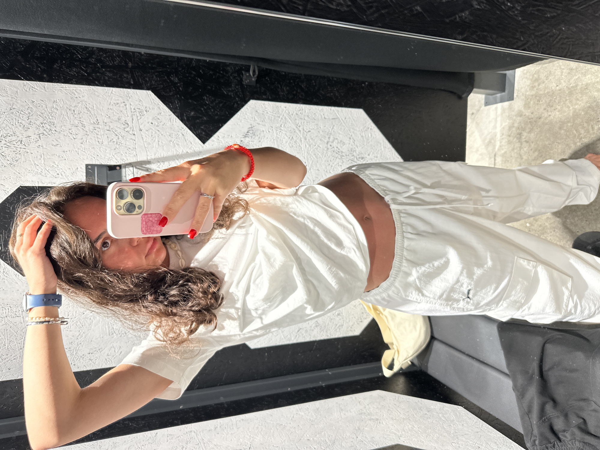 A person with long, curly brown hair is taking a selfie in a mirror. The person is wearing a white t-shirt and white sweatpants. A red bracelet is on their right wrist. The phone is pink. The background shows a black and white patterned surface. The location is likely an indoor space. It is not possible to confirm the location as Pattaya City, Thailand, without additional information.