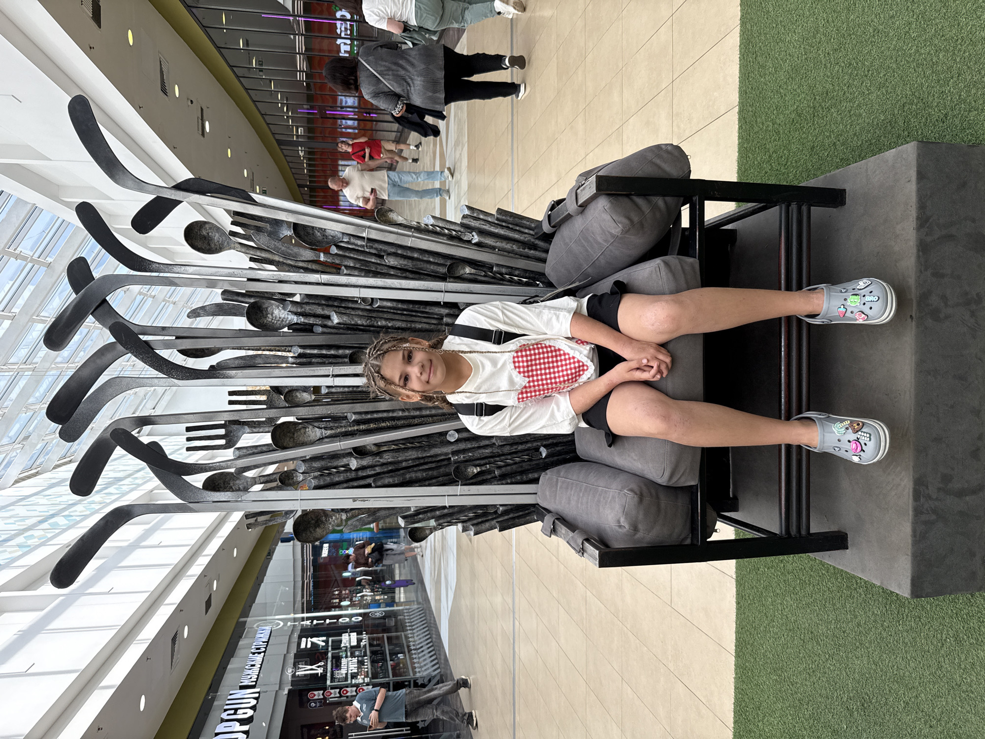 A young girl with braided hair sits on a large, dark gray sofa that is part of a sculpture made of cutlery. The sculpture resembles a throne, with tall, curved metal pieces sticking up behind and around the sofa. The girl wears a white t-shirt with a red and white checkered heart on the front, a black skirt, and gray Crocs with colorful charms. She is positioned in the foreground of the image.
The background shows a modern indoor shopping mall. The floor is tiled in a light beige color. To the left, there is a storefront with large windows displaying merchandise. A man in a blue polo shirt and jeans is walking by this storefront, looking at his phone. Further back, other shoppers are visible walking in the mall. The ceiling is high and features a glass roof with visible support beams, allowing natural light to enter. There is also artificial lighting from overhead fixtures. The area in front of the sofa is covered with artificial green grass. The scene is set in the city of Zhukovo, Russia.