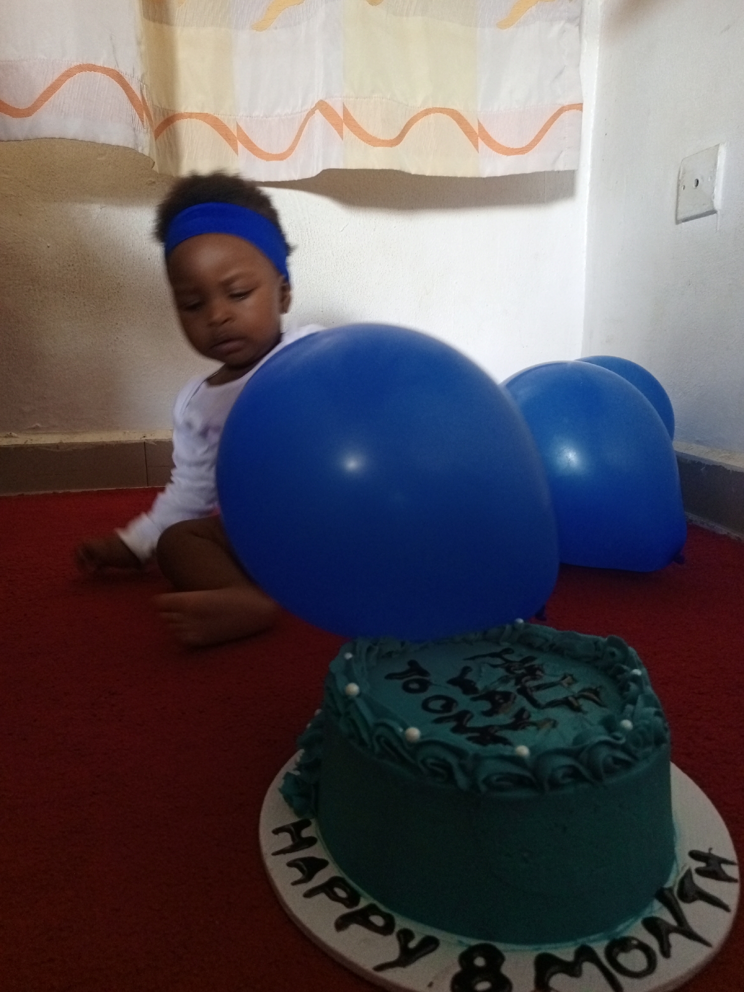 A baby wearing a blue headband sits on a red carpet in a corner of a room. The baby is looking slightly to the left, away from the camera. In front of the baby are two large blue balloons and a cake with "Happy 8 Month" written on its base. The walls are painted white, and a patterned curtain hangs in the background. The location is identified as Keffi, Nigeria. An electrical outlet is visible on the wall.