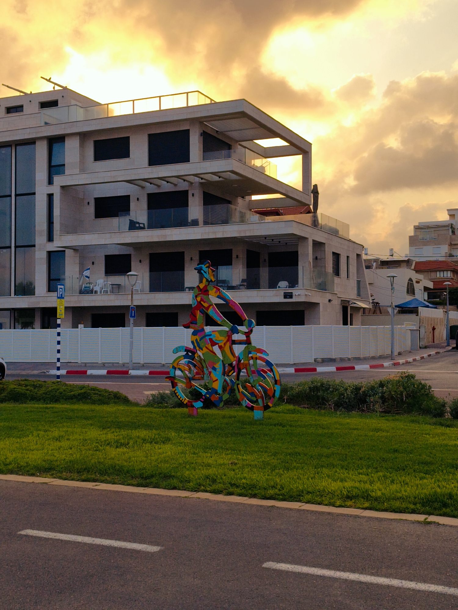 This is a photograph taken in the city of Nahariyya, Israel, depicting a modern apartment building situated near the coast. The sky above is filled with dramatic, brightly lit clouds, suggesting either sunrise or sunset. In the foreground, a vibrant, colorful sculpture of a person riding a bicycle is prominently displayed on a patch of green grass. The sculpture is abstract and geometric, composed of various bright colors like red, yellow, blue, and green.
The apartment building is contemporary in design, featuring multiple balconies, large windows, and a light-colored facade, possibly stone or stucco. Its architecture is characterized by clean lines and a modern aesthetic. In front of the building, a white fence with red and white striped bollards separates the property from the sidewalk and street. The street in the foreground has a white line marking a lane. To the right of the main building, other residential structures are visible, some with red-tiled roofs, indicating a densely populated urban or suburban area. The overall impression is a blend of modern urban living with artistic elements and the dramatic beauty of the sky.