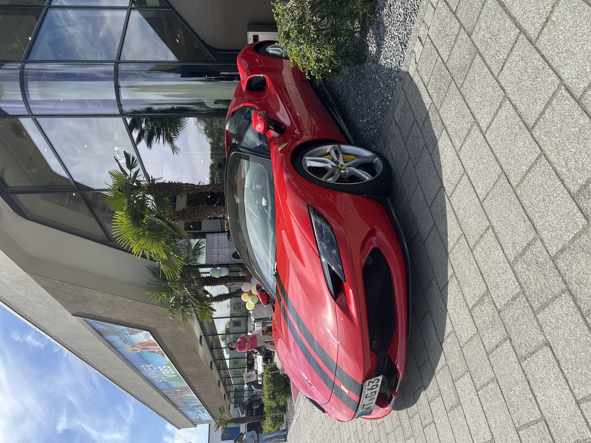 A red Ferrari SF90 Stradale is parked on a gray brick sidewalk in Jestetten, Germany. The car has black racing stripes and a license plate that reads "WT-G 663". A modern building with a glass facade is behind the car, reflecting palm trees and the sky. There are people, including one seated woman in a pink shirt, near a tent or sheltered area. The sidewalk abuts a narrow strip of gravel and greenery. Part of a sign with the words "SUMMER VIBES" is visible on the building's awning.