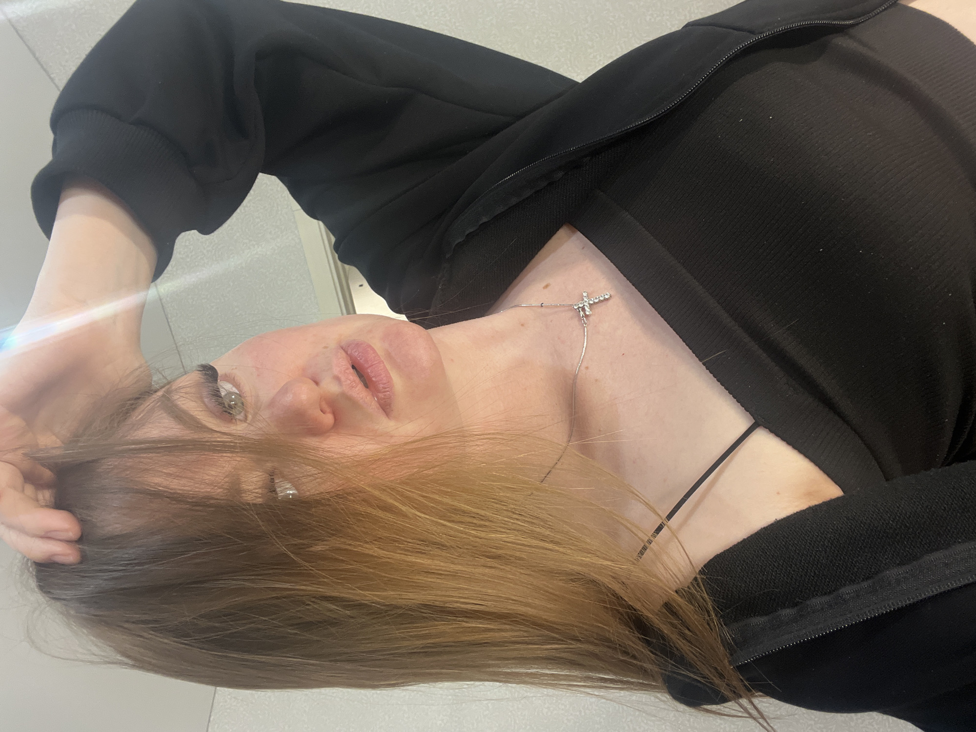 An indoor, medium shot captures a young woman from an overhead perspective, rotated 90 degrees counter-clockwise. Her head is positioned towards the left of the frame, with her body extending downwards. She has light brown to blonde hair, which cascades around her face and shoulder. Her light-colored eyes, possibly green or hazel, look directly towards the viewer with a neutral to slightly pensive expression. Her lips are naturally colored and slightly parted.
She is wearing a black, ribbed top, visible under a black zipped jacket or hoodie, which is partially open. A thin, silver-colored necklace with a small, sparkling cross pendant adorns her neck. A thin, dark strap, possibly from a bra or another necklace, is visible near her right shoulder (left in the image). Her left hand is raised, with fingers gently touching her hair near her temple.
The background is a light, indistinct surface, possibly a wall or ceiling, suggesting an indoor environment. Bright lighting illuminates the scene, and a minor lens flare is visible near her left hand. No specific activity beyond posing for the camera is evident. The image is set in Sevastopol, Ukraine, according to the provided context. There is no visible text in the image.