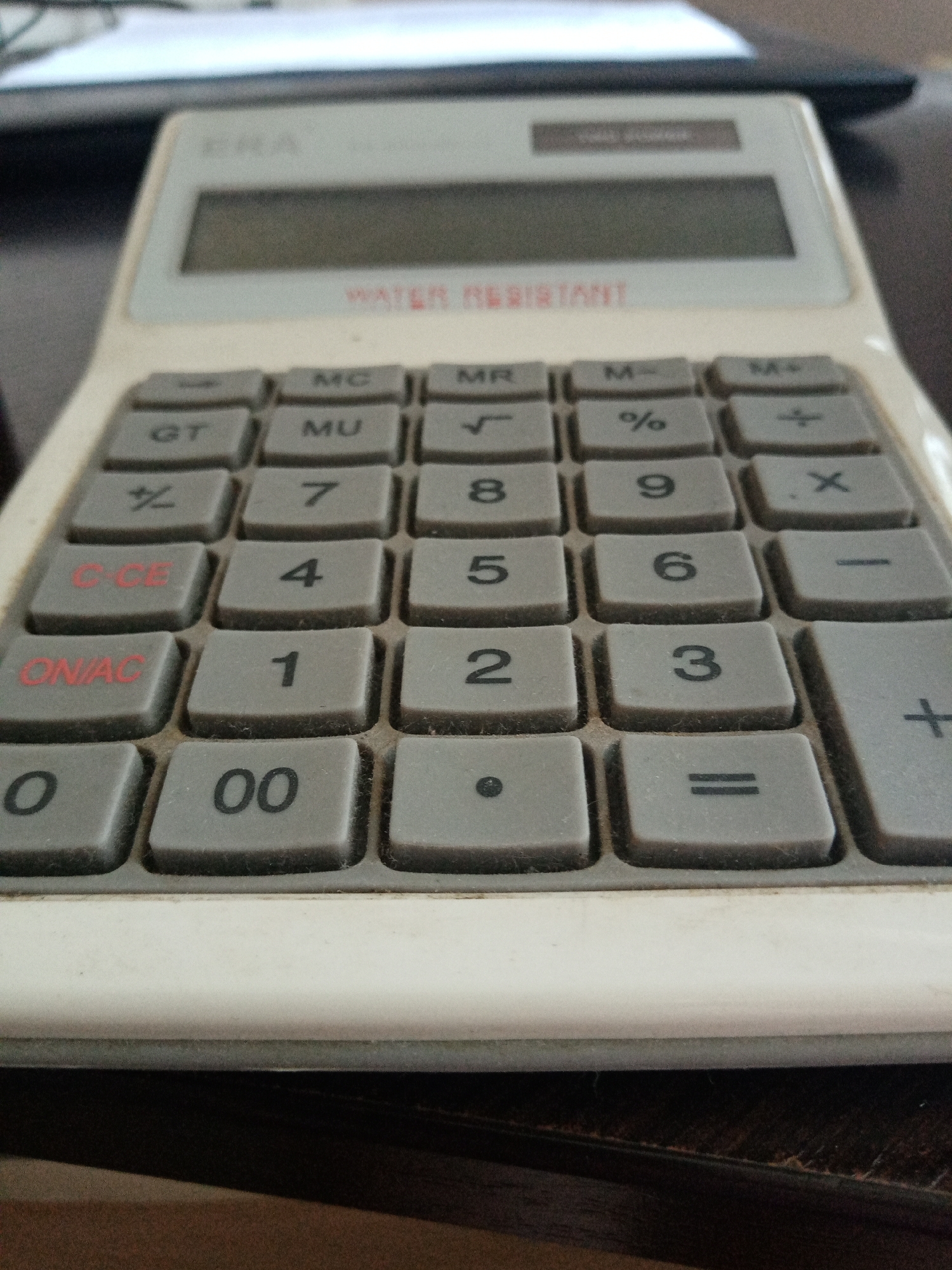 The image shows a close-up view of a well-used, off-white calculator. The calculator has gray rectangular buttons with black or red markings. The markings include digits, mathematical symbols, and abbreviations like "MC," "MR," "M-," "GT," "MU," "CE," and "ON/AC." Dust and grime are visible on the buttons and around the edges, indicating the calculator has been used frequently. The display window is located at the top and reads "WATER RESISTANT." The background is blurry, showing a dark wooden surface and what seems to be paperwork or a notepad behind the calculator. There is no indication of location in Elektrostal, Russia.