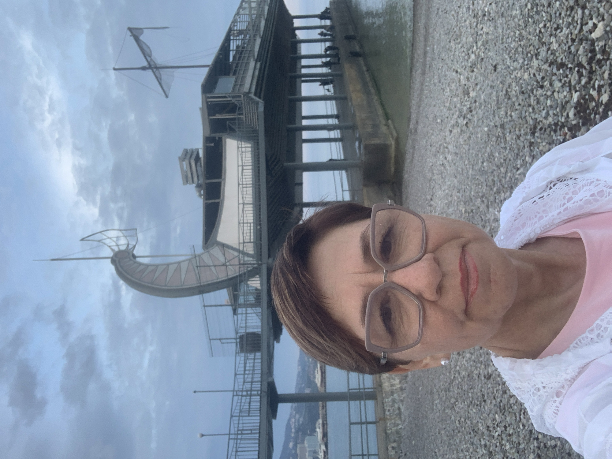 A person wearing glasses, a pink shirt, and a white lace garment is visible in the foreground. The backdrop reveals a cloudy sky and a stone beach. The "Floating Sculpture of Thessaloniki" is a prominent architectural feature, with a boat-like structure supported by a steel platform. Below, a glimpse of a cityscape or coastal area is present. The image is rotated to the left. The location is near the "Floating Sculpture of Thessaloniki" in Thessaloniki, Greece.