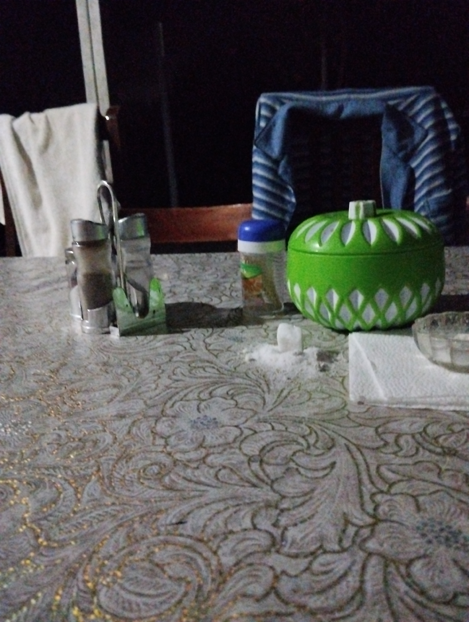The image depicts a close-up, low-light, indoor scene, likely on a table covered with a patterned tablecloth. In the foreground, a salt shaker and a pepper shaker are visible, held in a metallic stand. To the right of the shakers, a decorative green container with a lid, resembling a sugar bowl or candy dish, is present. A small pile of white granules, likely salt, is scattered on the tablecloth near the green container. To the right of the green container, a white paper napkin and a glass bowl can be seen. In the background, a striped blue and white fabric, possibly clothing or a blanket, is draped over a dark object. A white towel hangs on the left side of the frame. The overall lighting suggests it is nighttime or a dimly lit room. There is no indication of the specific city of Müqtədir, Azerbaijan.