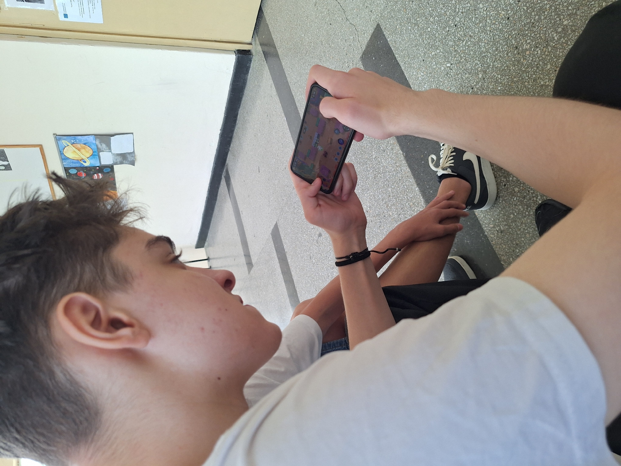 In Varna, Bulgaria, a young person is seated on the floor, engrossed in a mobile game on their smartphone. They are wearing a white t-shirt and dark shorts, with a black woven bracelet on their left wrist. Their right hand holds the phone, and their left thumb is positioned to interact with the screen. Another person is partially visible beside them, also seated. The background shows a lightly colored wall with some artwork and a door. The floor is tiled with a speckled pattern and separated by dark lines.