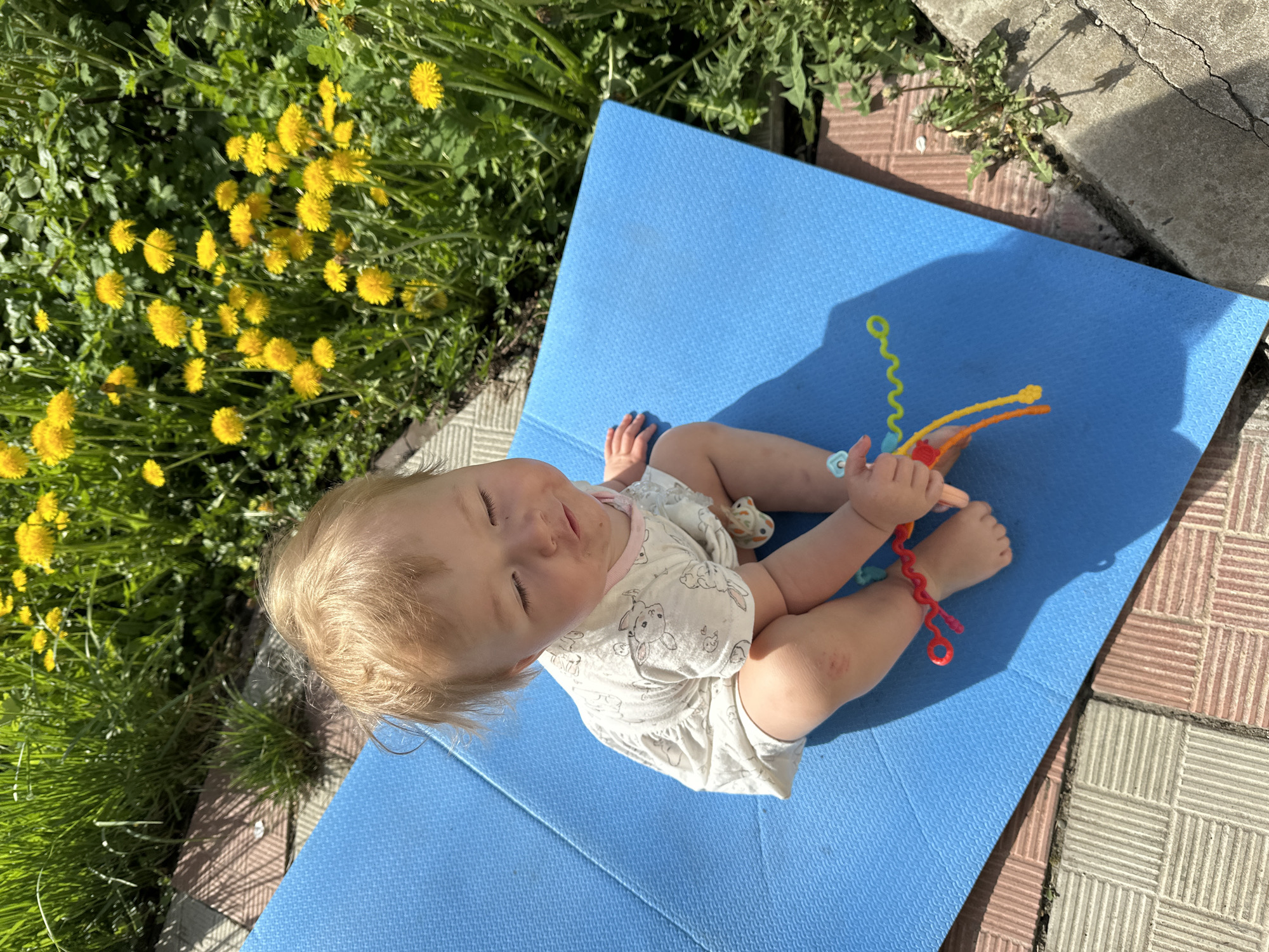 A baby sits on a folded blue mat outdoors, possibly in Nizhny Novgorod, Russia. The child is fair-skinned with blonde hair and is wearing a white romper with bunny prints. The baby holds a colorful toy made of fuzzy, flexible wires. A patch of dandelions grows nearby. The mat is placed on tiled patio stones next to a concrete edge. The scene is brightly lit, indicating daytime.