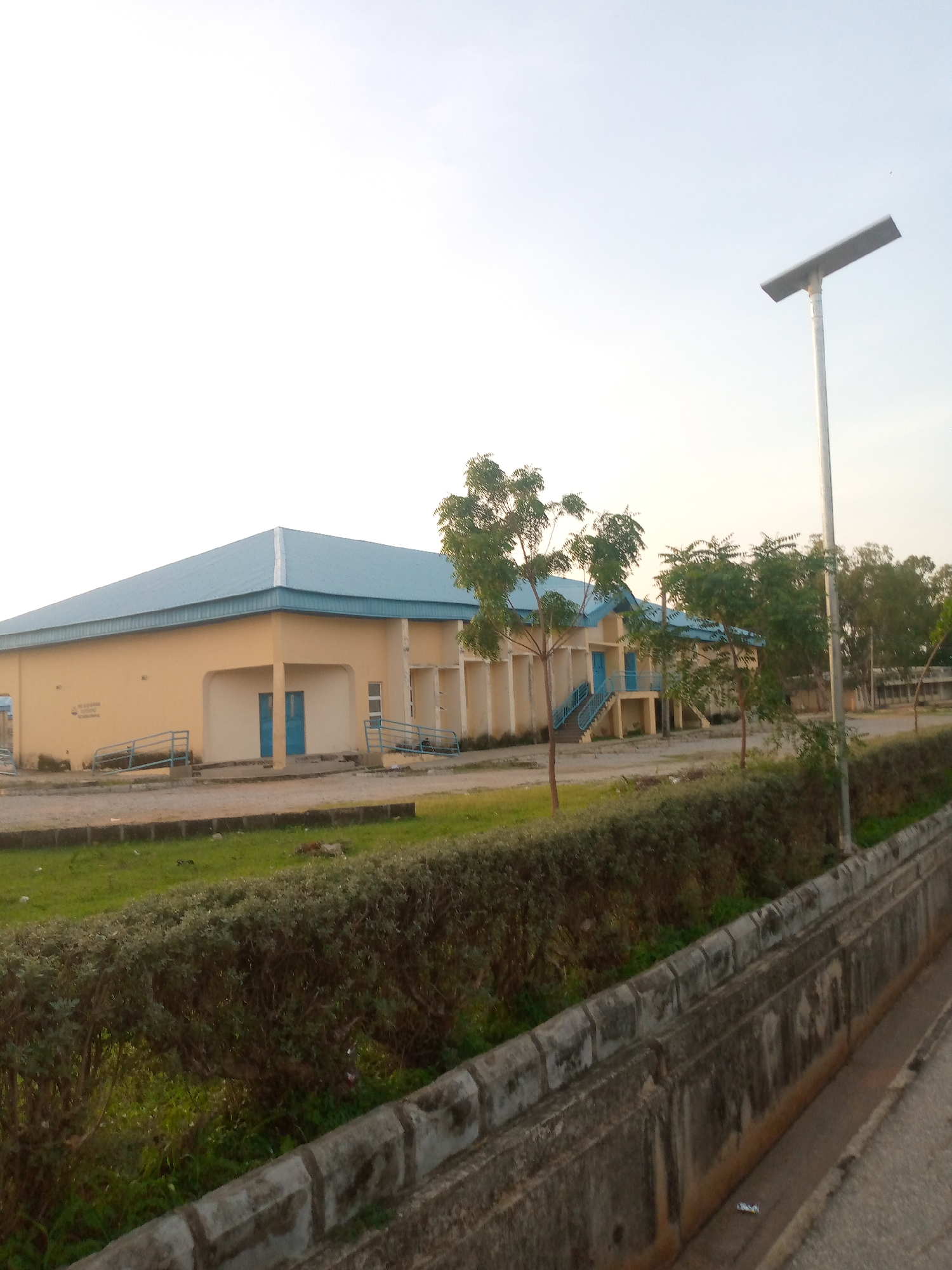 The image shows a low-rise, light yellow building with a light blue roof, situated in Jalingo, Nigeria. The building features several blue doors and windows, with ramps leading up to some of the entrances. The structure appears to be a school or administrative building, with trees and manicured bushes surrounding it. A solar-powered street light stands tall to the right of the building, indicating a modern infrastructure. The scene is set against a clear, bright sky, suggesting daytime. The ground is a mixture of paved areas and grassy patches, with a concrete drainage channel running along the foreground. No people are visible in the frame, lending a sense of quietude to the location.