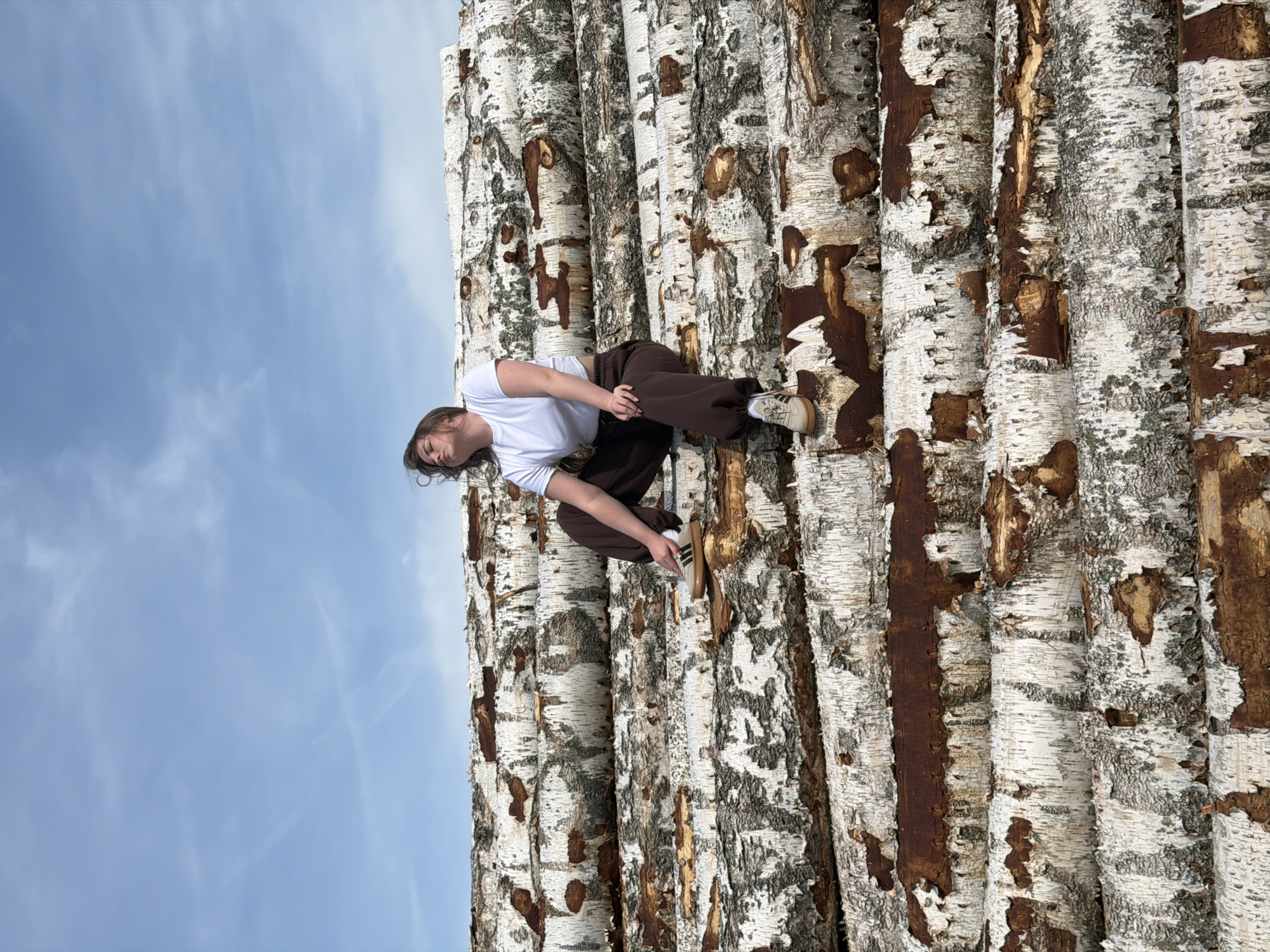 A woman is lying on a stack of birch logs against a blue sky. She is wearing a white shirt, brown pants, and brown shoes with white stripes. The logs have white and brown bark. The location is not specified. The scene is outdoors.