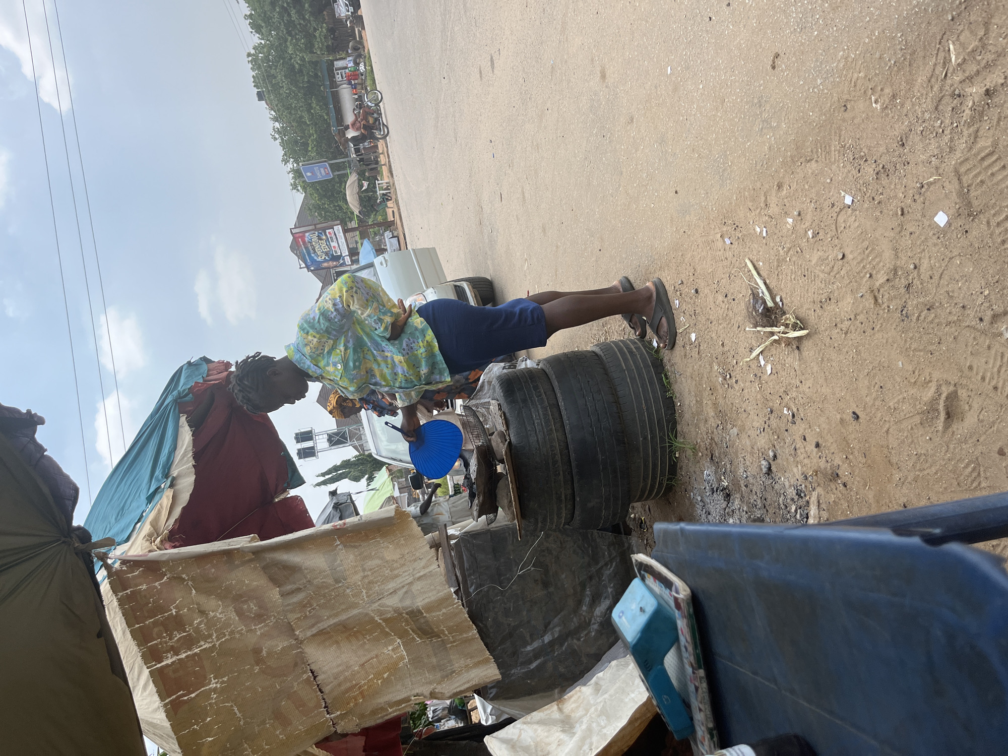 A person with dark hair is standing and holding a blue fan. They are wearing a colorful shirt and dark shorts. The person is near a stack of tires. In the background, there are tents or makeshift shelters made of tarps and fabric. There are also cars and buildings visible, suggesting an outdoor, possibly urban or roadside setting. The ground is sandy and dusty, with some debris scattered around. A white vehicle is parked behind the person and tires. The sky is overcast. This scene appears to be set in Abuja, Nigeria.