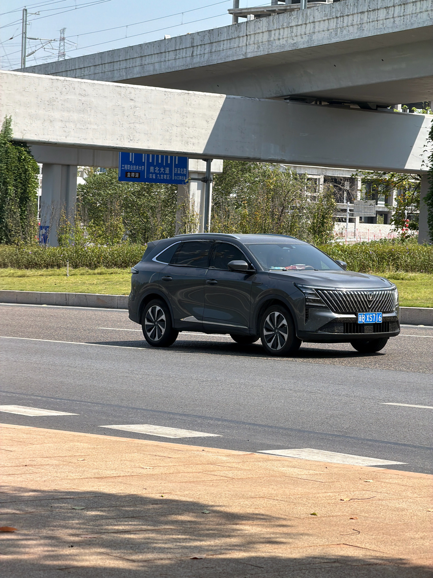 An exterior, medium shot captures a dark gray SUV driving on a paved road. The vehicle is positioned on the right side of the frame and is moving forward. The SUV has a visible license plate that reads "B XS716". Behind the car, a green lawn stretches out, bordered by a concrete curb and then a road. In the background, a large, elevated concrete structure, likely an overpass or part of a highway, dominates the scene. Beneath this structure, signage with Chinese characters is visible, indicating directions and place names within the city of Jiangjin, China. The sky is clear and blue, suggesting daytime. The foreground shows a textured pedestrian walkway with a light brown hue.