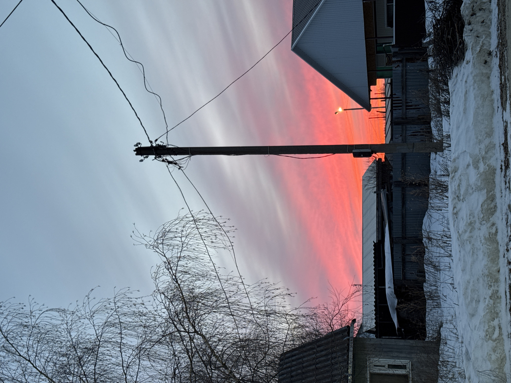 The image depicts a street scene in Vysokiy, Russia, during a vibrant sunset. The sky is striated with shades of red and white. A utility pole stands prominently in the foreground, with electrical wires stretching across the frame. Bare tree branches are visible below the pole. To the right, a building with a gray roof and siding is partially obscured by a snowdrift. A streetlight is illuminated next to the building.