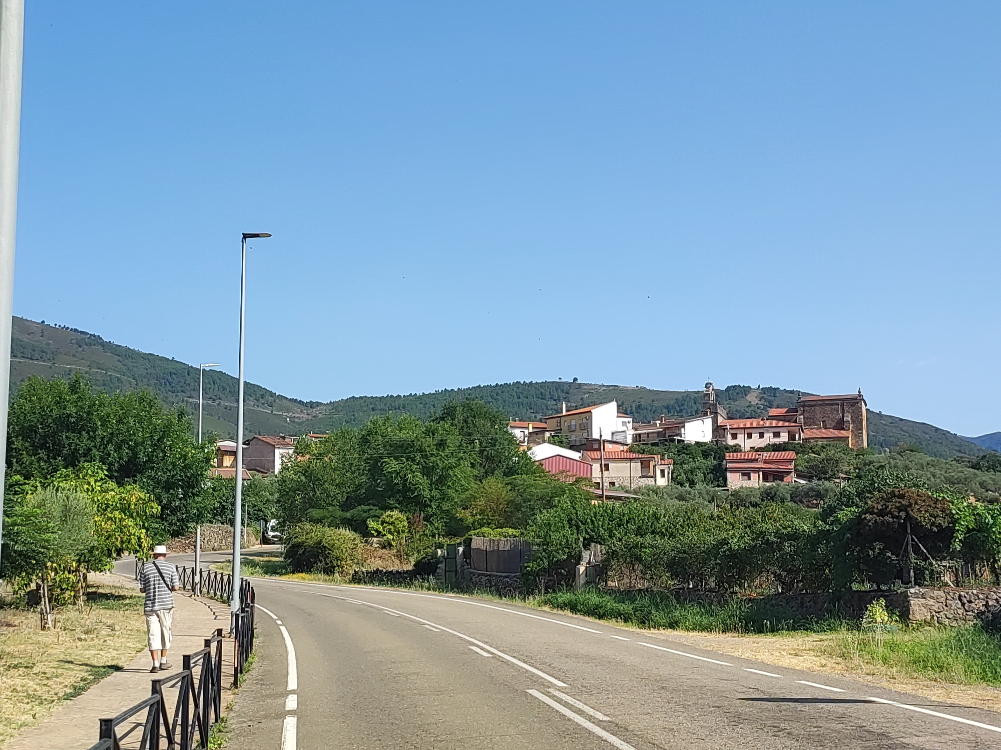 The image depicts a sunny day in Cadalso, Spain, showing a rural road with a man walking on the sidewalk. The road curves to the right, and a black metal fence runs along the sidewalk. Behind the fence, there are trees and vegetation. In the background, a village is nestled on a hillside, with houses featuring terracotta roofs. The sky is clear and blue. The man is wearing a striped shirt and light-colored shorts, and he is walking away from the camera. A street lamp is visible on the left side of the image. The overall scene suggests a quiet, residential area in a hilly landscape.