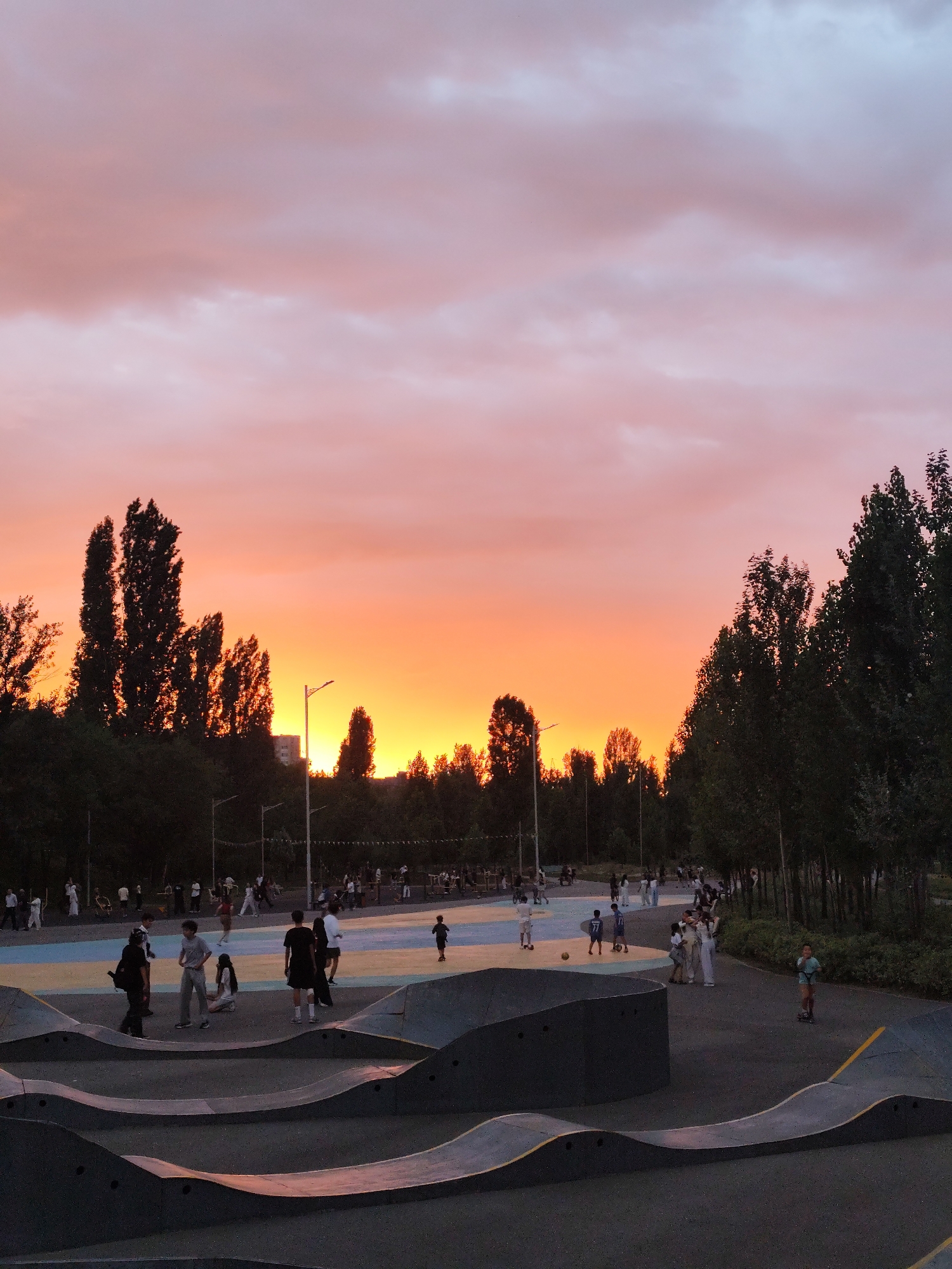 The image depicts a vibrant sunset over a skate park in Almaty, Kazakhstan. The sky is ablaze with hues of orange, pink, and purple, casting a warm glow over the scene. In the foreground, a series of undulating concrete ramps form the skate park, with a few individuals scattered throughout the area. Some are engaged in activities on the ramps, while others are standing and observing.
In the middle ground, a colorful paved area with blue, yellow, and white sections serves as a basketball court or a similar recreational space. More people are present here, some playing with a soccer ball, while others are gathered in groups. The background is lined with tall, dark green trees, silhouetted against the bright sky. A few streetlights are visible, indicating the urban setting. The overall atmosphere is one of a lively evening gathering, with people enjoying the public space as the day transitions into night.