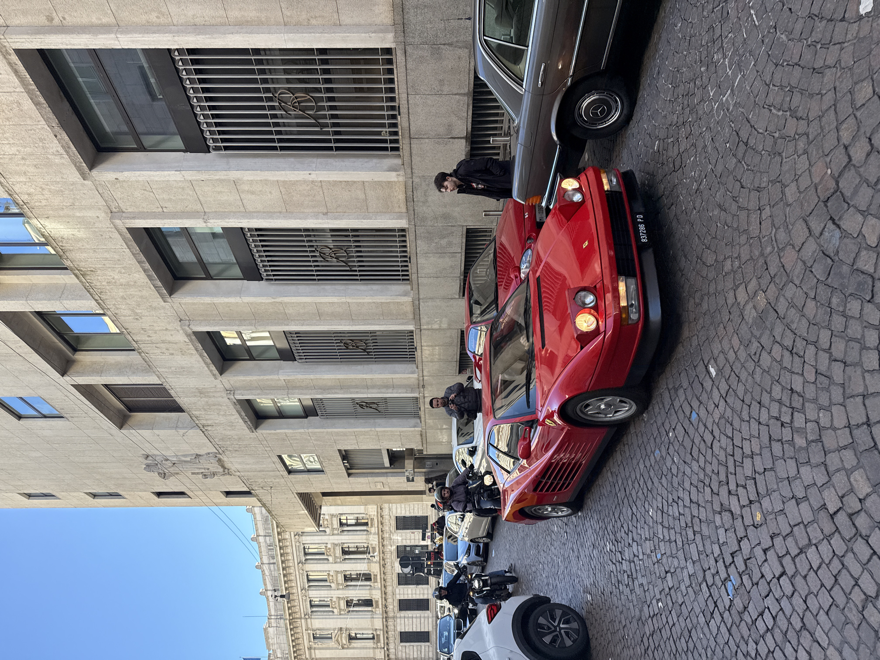 This image depicts a bustling daytime street scene in Milan, Italy, specifically in the vicinity of Piazza della Scala, characterized by a mix of historic architecture and urban traffic. The foreground features a cobblestone street, paved with dark gray granite blocks arranged in an arc pattern.
To the right, a multi-story building of modern-classical design dominates the frame. Its facade is composed of light-colored stone blocks, with regularly spaced rectangular windows framed in dark material. Several ground-level and first-floor windows are protected by vertical metal grilles, each featuring a stylized "B" initial in the center, identifying it as the former Banca Commerciale Italiana headquarters (now Intesa Sanpaolo). Higher up, subtle stone relief carvings are visible on the facade.
In the mid-ground, a vibrant red Ferrari Testarossa is the central subject, facing slightly left with its pop-up headlights illuminated. Its Italian license plate, "837286 PD," is clearly visible on the front bumper. To the immediate right of the Ferrari, a dark brown vintage Mercedes-Benz sedan is positioned, with its chrome trim and a Mercedes-Benz branded hubcap visible. A young man with dark hair, wearing a black jacket, stands on the cobblestones between the Ferrari and the Mercedes, looking left. Behind the Ferrari, a second red sports car is partially visible, along with a man in a dark puffer jacket holding a camera or smartphone, actively photographing the Ferrari.
On the left side of the street, a white SUV's rear section is visible in the foreground. Two men on motorcycles navigate the street; both wear helmets. One rider in a dark jacket and blue jeans is on a black motorcycle, while another in a gray hoodie and dark jacket is on a dark motorcycle. In the background, numerous other vehicles, including a white BMW SUV, a blue hatchback, and additional sedans, indicate moderate urban traffic. Further back, an ornate classical building, resembling the Palazzo del Credito Italiano on Piazza Cordusio, is discernible with its detailed facade and arched windows. Pedestrians are sparsely visible on sidewalks in the distant background. The sky above is clear and blue, suggesting fair weather.