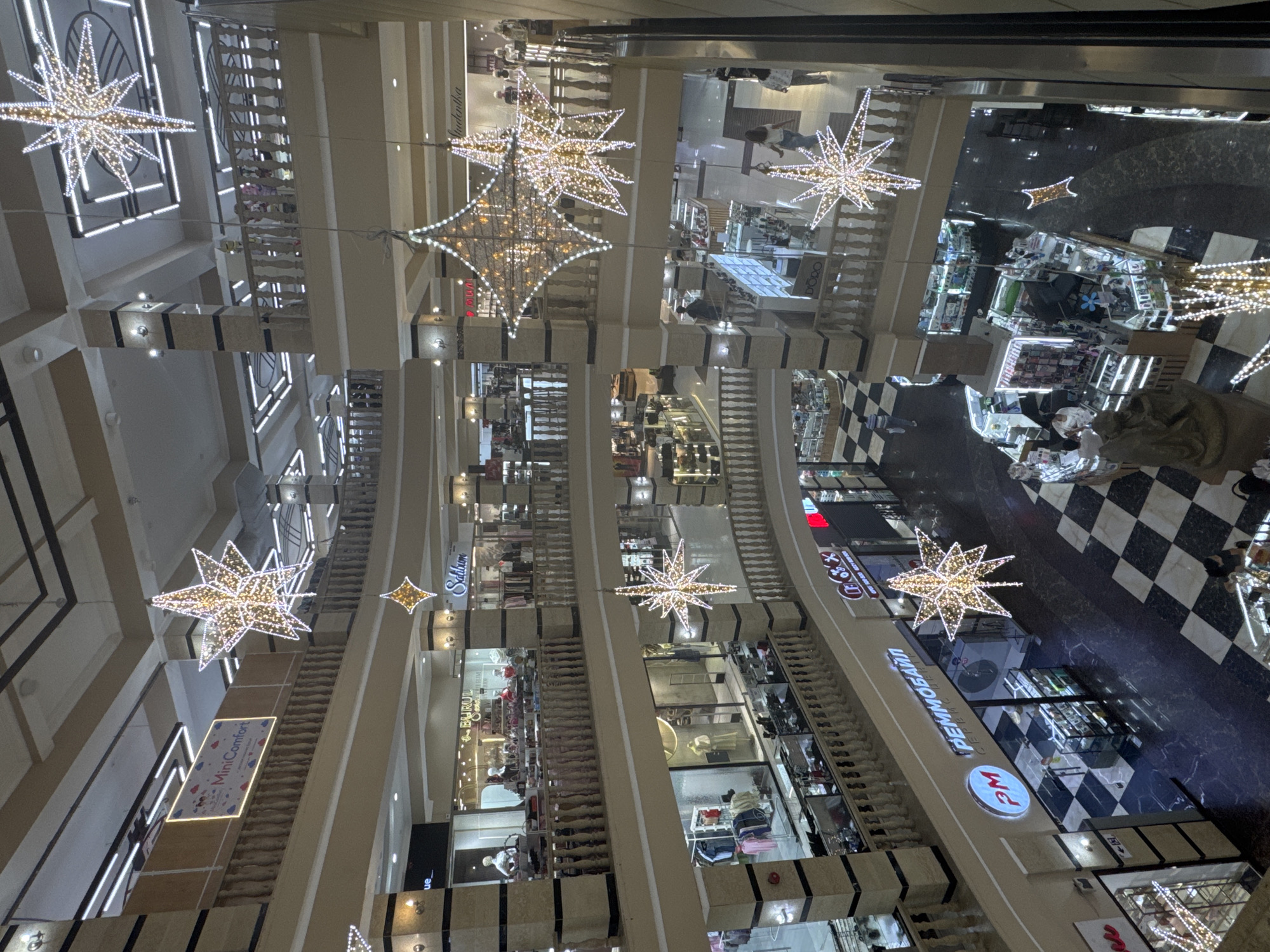This is an interior shot of a multi-level shopping mall, likely in Bishkek City, Kyrgyzstan, adorned with festive star-shaped light decorations. The mall features several floors with open walkways and balconies overlooking the central atrium. Various shops with glass storefronts display merchandise, including clothing and possibly electronics. People are visible on different levels, browsing or walking through the mall. The floors are a mix of checkered black and white tiles and darker marble. The lighting is predominantly artificial, with the bright star decorations creating a festive atmosphere.