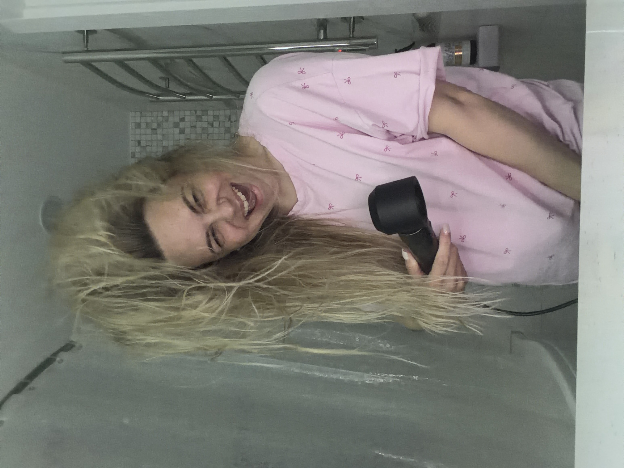 A woman is in a shower stall. She is holding a hair dryer and smiling. She has long blonde hair that is wet. She is wearing a pink pajama top with bow designs. The shower stall has a shower curtain that is drawn, partially obscuring the woman's lower body. A towel rack with towels on it is mounted on the wall of the shower stall, along with some white and grey tile. The location is not known to be in Votkinsk, Russia.