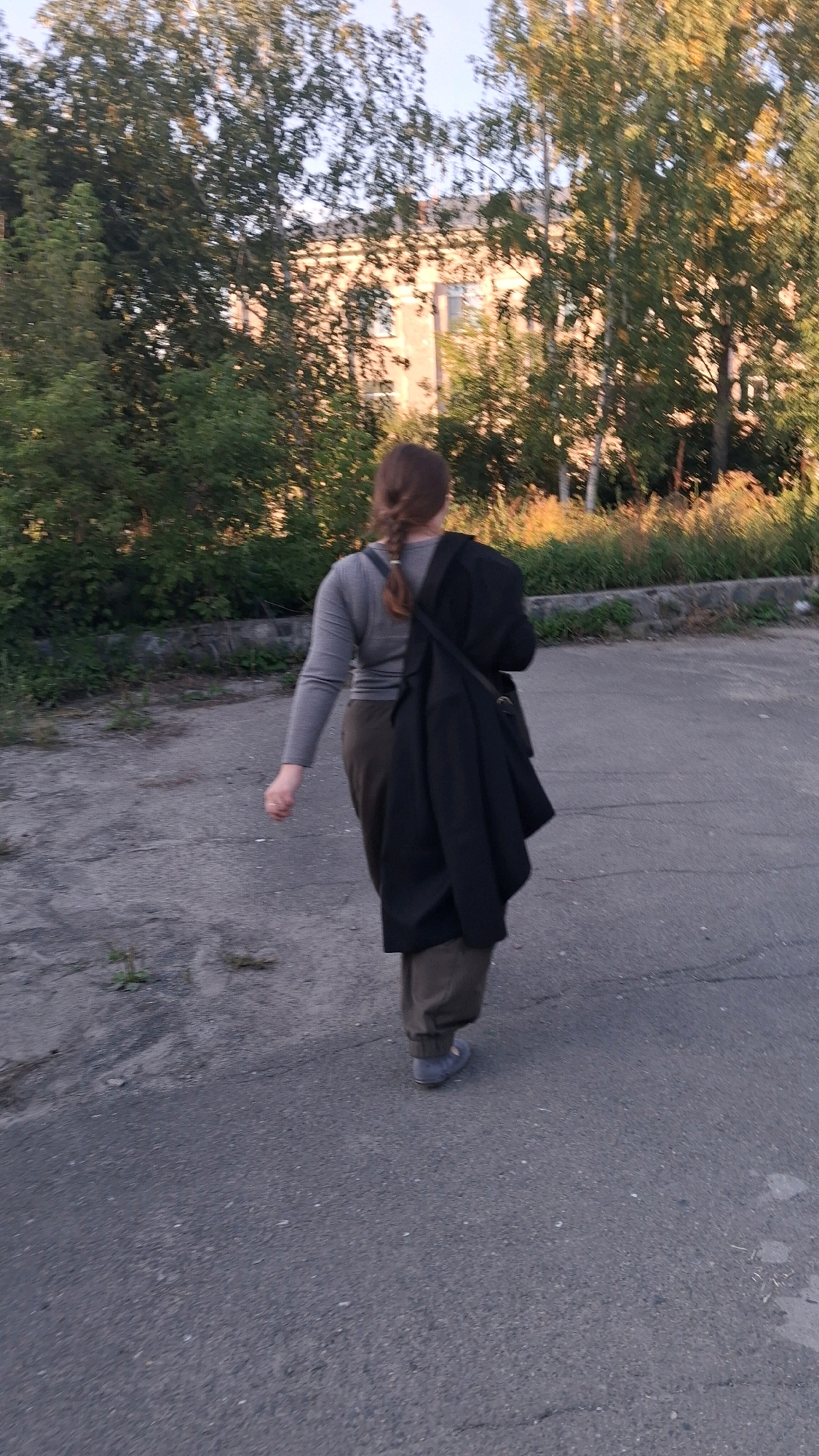 The image depicts a person with their back to the camera, walking away on an asphalt surface. The person is wearing a grey long-sleeved shirt, dark pants, and grey shoes. A dark coat is draped over their left shoulder. Their hair is styled in a braid. The background features trees with foliage, suggesting an outdoor setting. Behind the trees, a light-colored building with windows is visible. The overall environment appears to be a somewhat unkempt urban or suburban area. The lighting suggests it is either early morning or late afternoon. The image does not provide any specific landmarks or indicators to pinpoint a location within Novosibirsk, Russia.