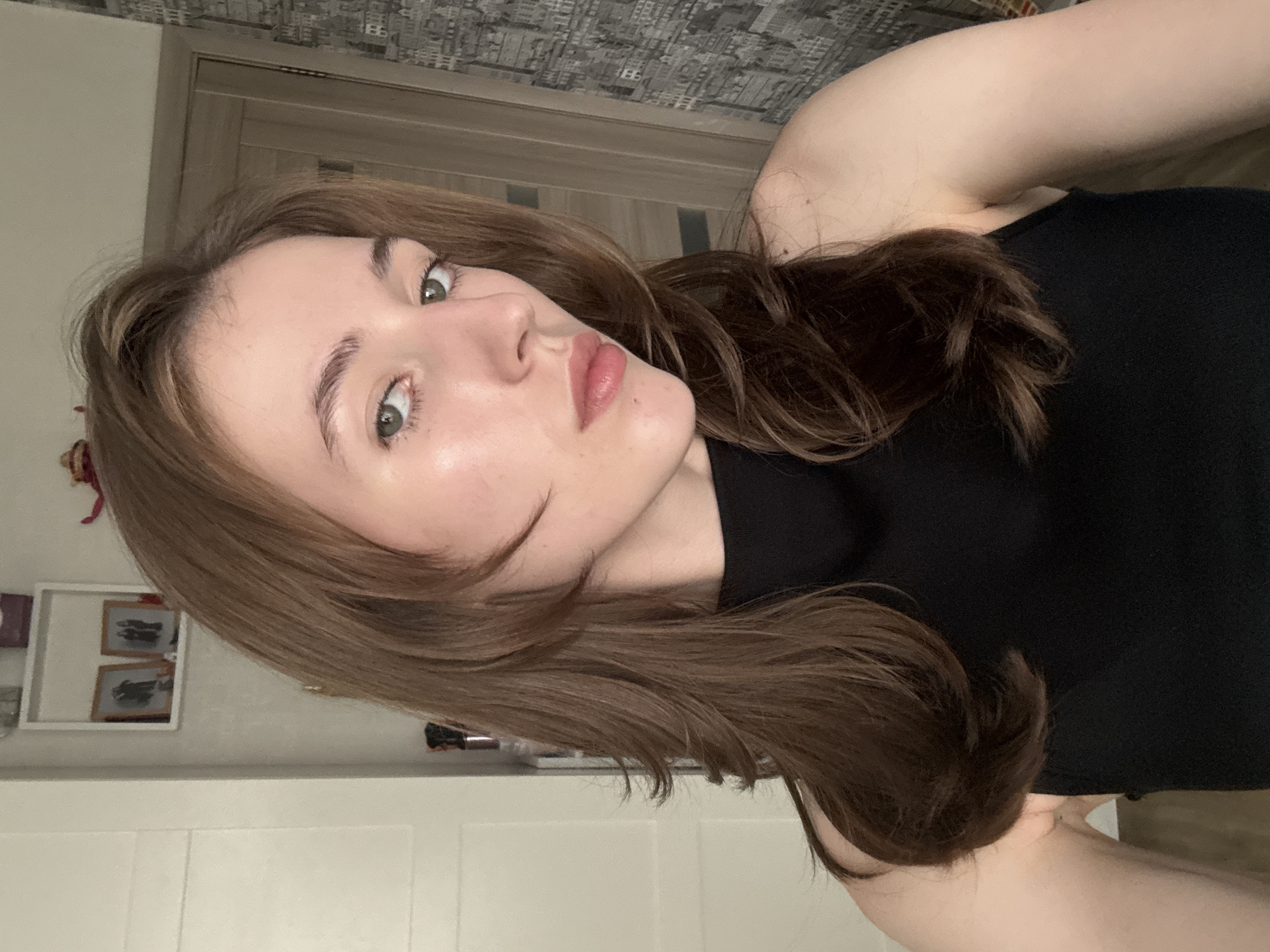 A young Caucasian woman with medium-length brown hair and green eyes is captured in a close-up, waist-up selfie. She is wearing a black sleeveless top. The background reveals a room with a white door on the left and a patterned wallpapered wall on the right, possibly indicating an interior in a residential building. A narrow shelf with two framed pictures is visible on the wall above her left shoulder. The lighting suggests an indoor setting, possibly in the city of Perm, Russia.