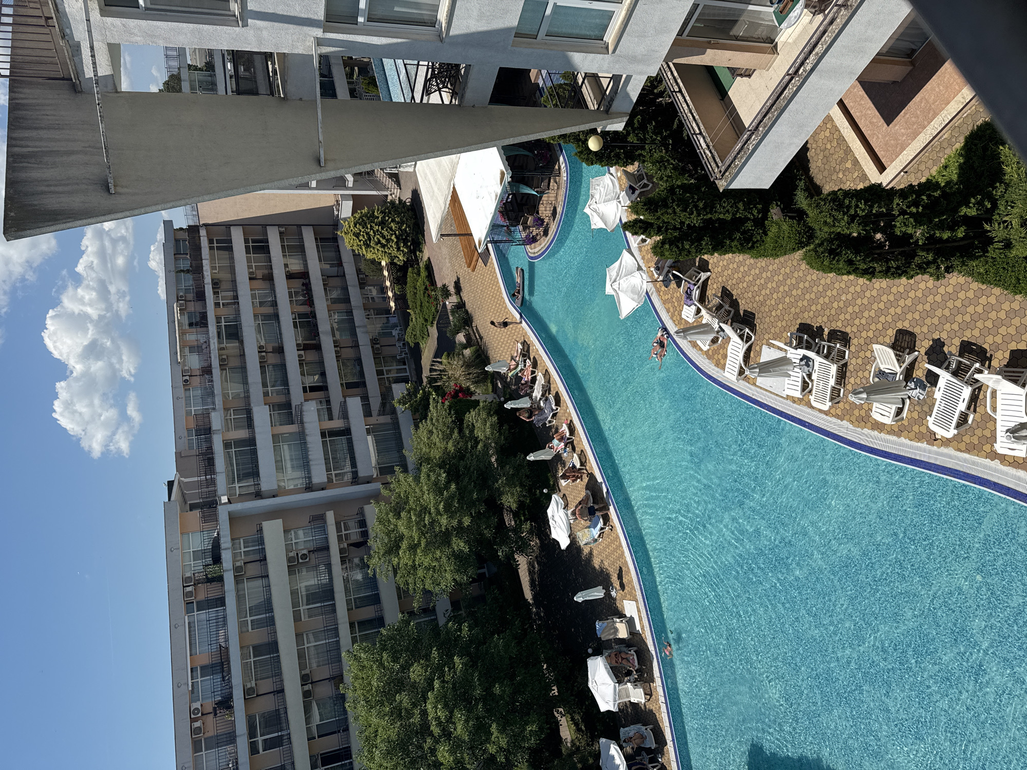 This is a high-angle photograph of an outdoor swimming pool and surrounding hotel buildings on a sunny day. The pool, filled with clear turquoise water, curves in a large "S" shape. Several people are visible in and around the pool. One person is swimming, and two others are lying on inflatable rafts. Around the pool, lounge chairs and white umbrellas are set up on a paved patio. A few people are seated on the lounge chairs.
In the background, there are multi-story hotel buildings with balconies and numerous windows. Trees and landscaping are also present, adding greenery to the scene. A covered outdoor bar or seating area is situated adjacent to the pool. The sky is bright blue with a few scattered white clouds. The overall impression is of a resort or hotel complex, likely in a warm climate, given the presence of the pool and people in swimwear. This scene is located in Nesebar, Bulgaria.