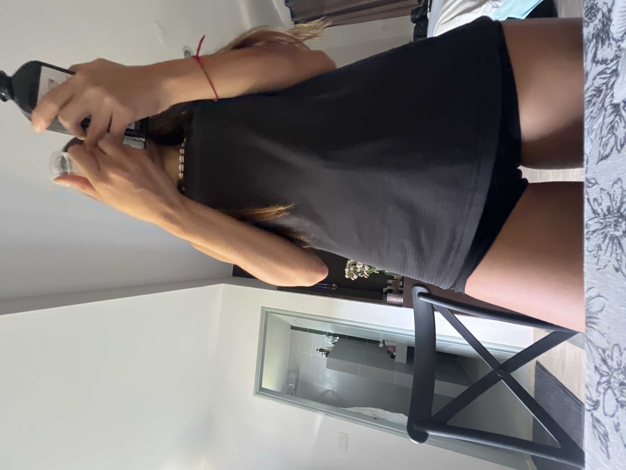 The image shows a person pouring liquid from a dark bottle into a small, clear cup. The person is wearing a black top and shorts. They also have a bracelet on their left wrist and a necklace.
The background includes a bathroom with white tiles and various toiletries, indicating a domestic setting. A black chair is visible in the foreground. The location may be in บ้านบางรักษ์, Thailand.