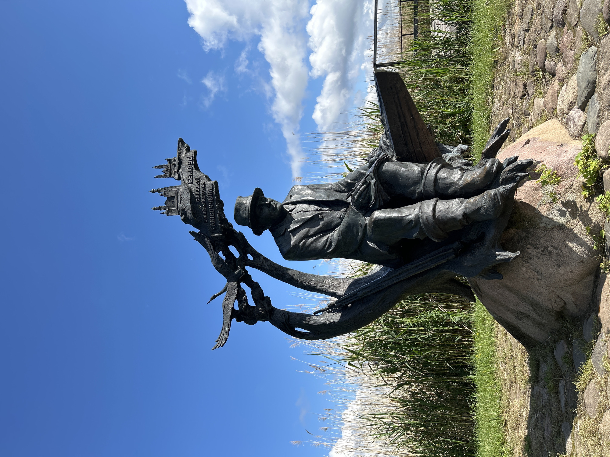 A bronze statue of a man sitting on what appears to be a wooden bench or plank is the central subject of the image. The man is wearing a hat, a suit jacket, and trousers tucked into boots. He is holding something in his hands, possibly a book or papers, and appears to be looking down at it. Behind him and to his left, a stylized bronze tree trunk curves upwards, supporting a depiction of a city skyline and what looks like a bird in flight. Text is inscribed on the city skyline portion, with the word "БЕЛАРУСЬ" clearly visible, indicating a connection to Belarus. The statue is positioned on a large rock, surrounded by cobblestones and grass. In the background, tall reeds or dry grasses grow, and beyond that, a wooden fence and a cloudy blue sky are visible. The scene is set outdoors, likely in a park or natural area, given the vegetation and open sky. The statue is located in the village of Azaretski, within the Azaretski Village Council, Belarus.