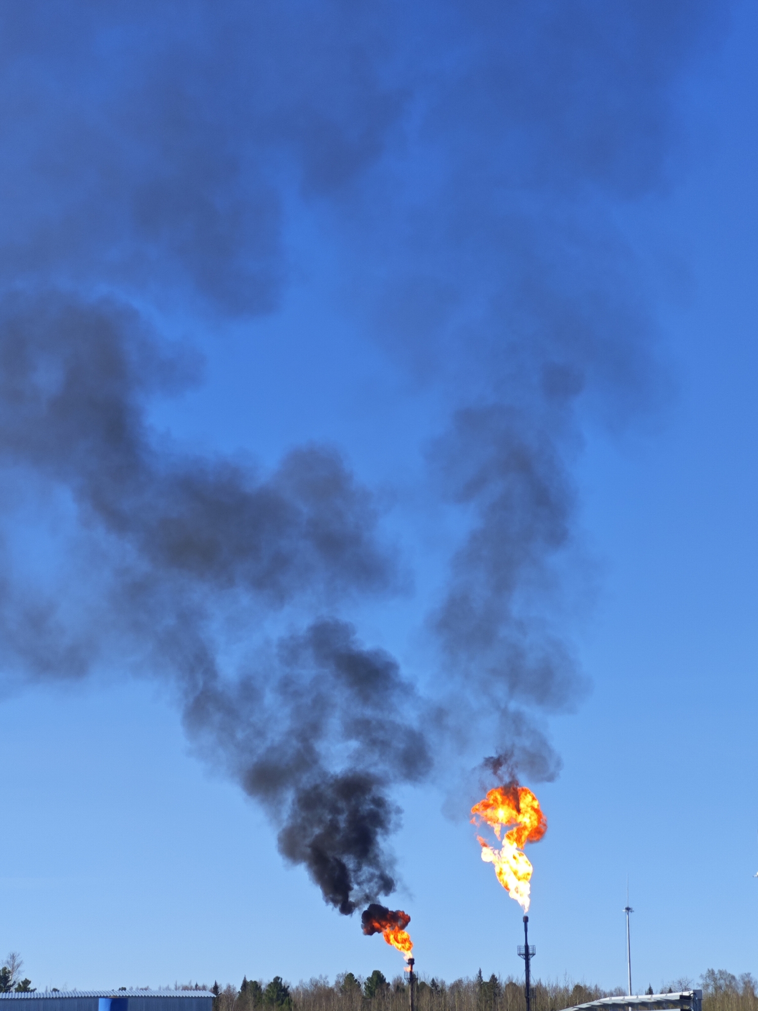 The image depicts two gas flares burning against a clear blue sky, producing significant plumes of black smoke. The flares are located behind a line of trees and a low, industrial-looking building. Several utility poles are visible. The location is within the city of Unknown, Russia. The scene suggests industrial activity, likely related to oil or gas production.