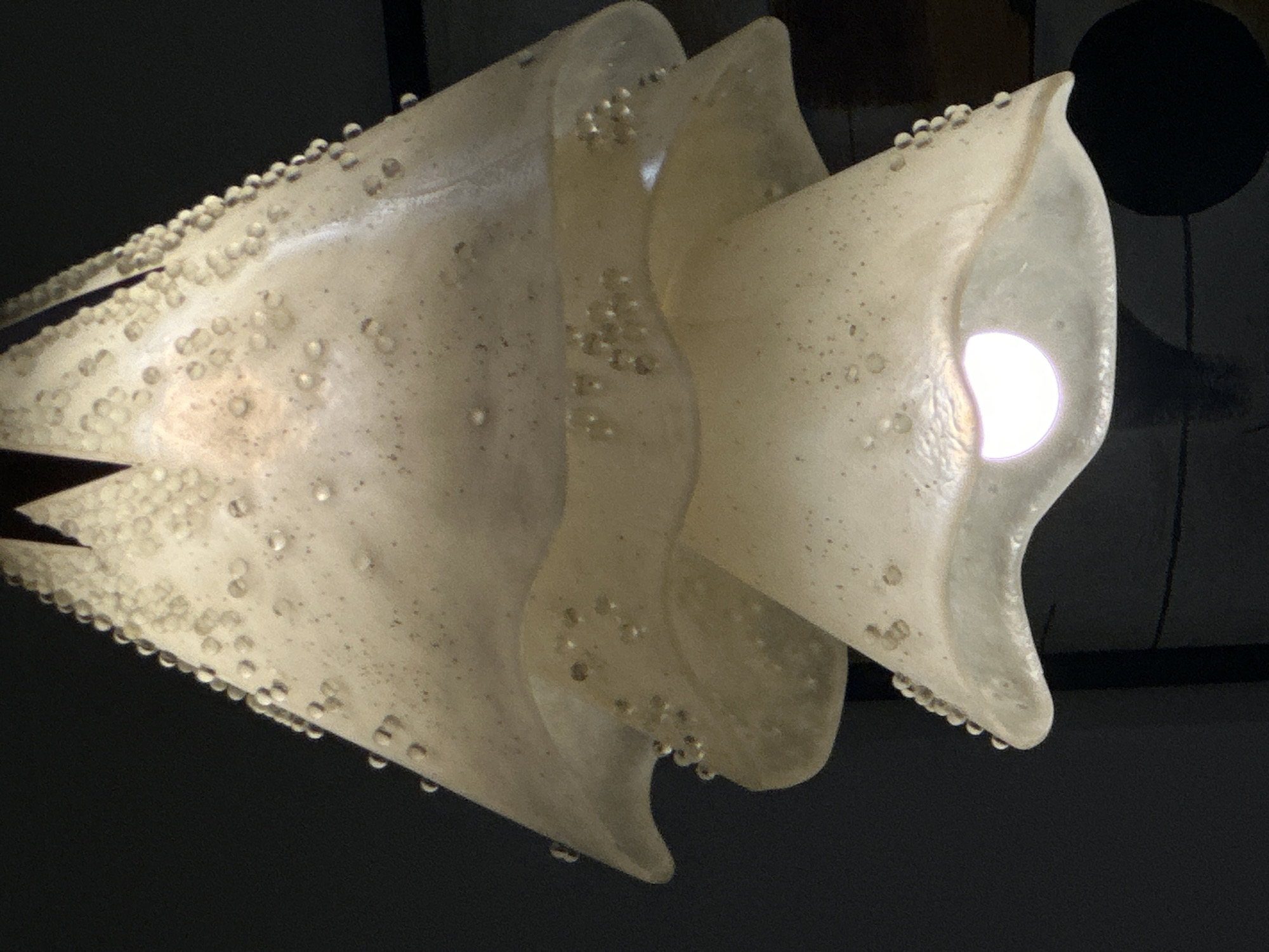 The image shows a close-up of a decorative light fixture. The fixture is composed of three overlapping, translucent, conical shades that are white or off-white in color. Each shade is decorated with numerous small, clear, spherical beads clustered along the edges and scattered across the surfaces. The beads give the fixture a textured, almost bubbly appearance. The bottom-most shade has a lightbulb emitting a bright white light from within. The background is dark and out of focus, suggesting an indoor environment, possibly at night or in a dimly lit room. There is no indication of people, actions, or specific environmental details that would allow for pinpointing a location in San Bernardino Tlaxcalancingo, Mexico.