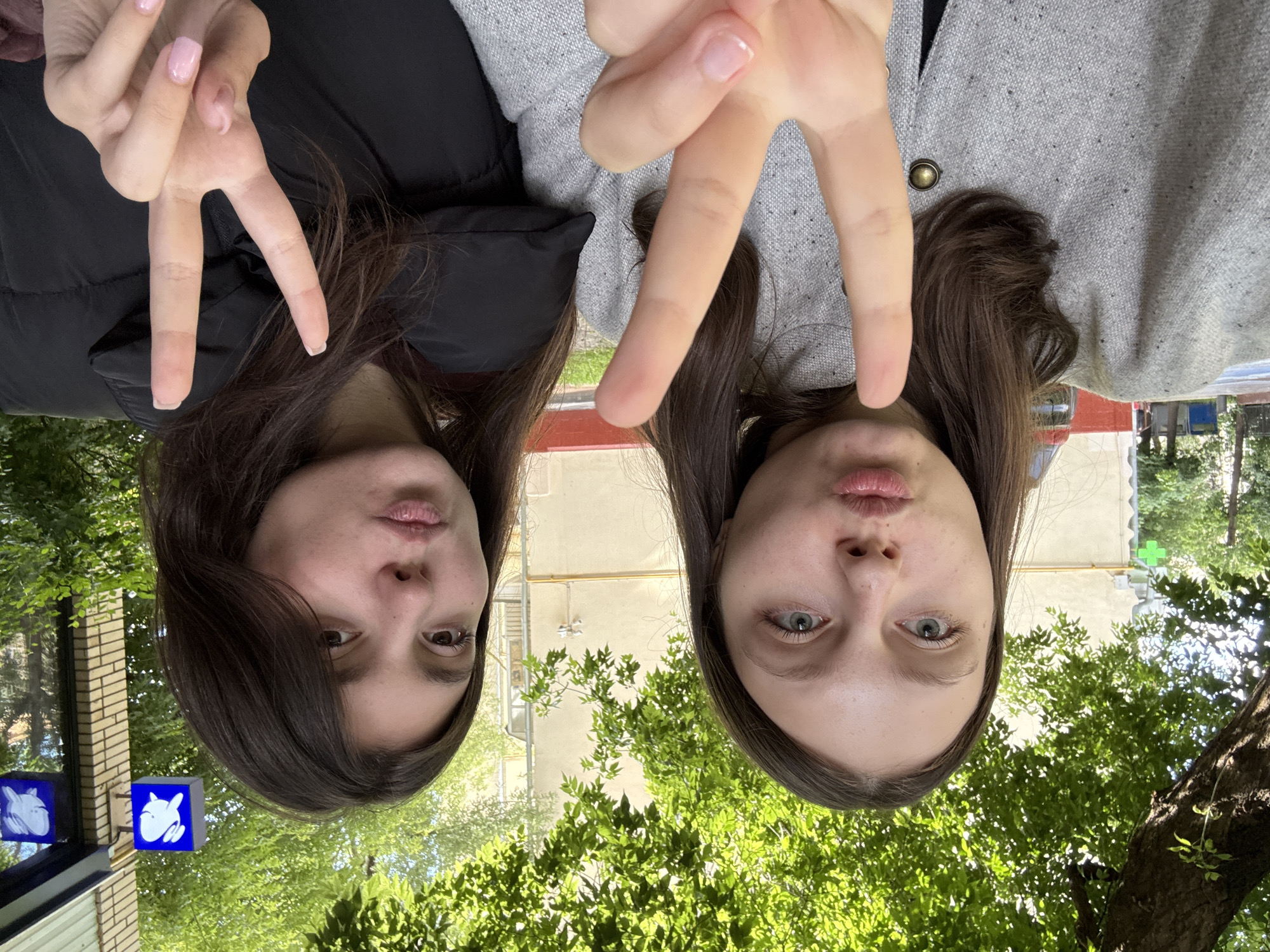 Two young women are posing for a selfie outdoors. The woman on the left is wearing a grey textured jacket with a brass button, and the woman on the right is wearing a black puffer jacket. Both women are making a "peace sign" gesture with their fingers and puckering their lips. Behind them, a building with yellow-beige siding is visible, along with trees and a portion of a street. In the upper right corner, two blue square signs with white rabbit illustrations are mounted on a brick wall, indicating the location is likely a children's cafe or store. These signs are characteristic of "Rabbit" cafes in Moscow, Russia, which are known for their themed decor and often feature live rabbits. The presence of the rabbit signs strongly suggests the photo was taken near one of these establishments in Moscow.