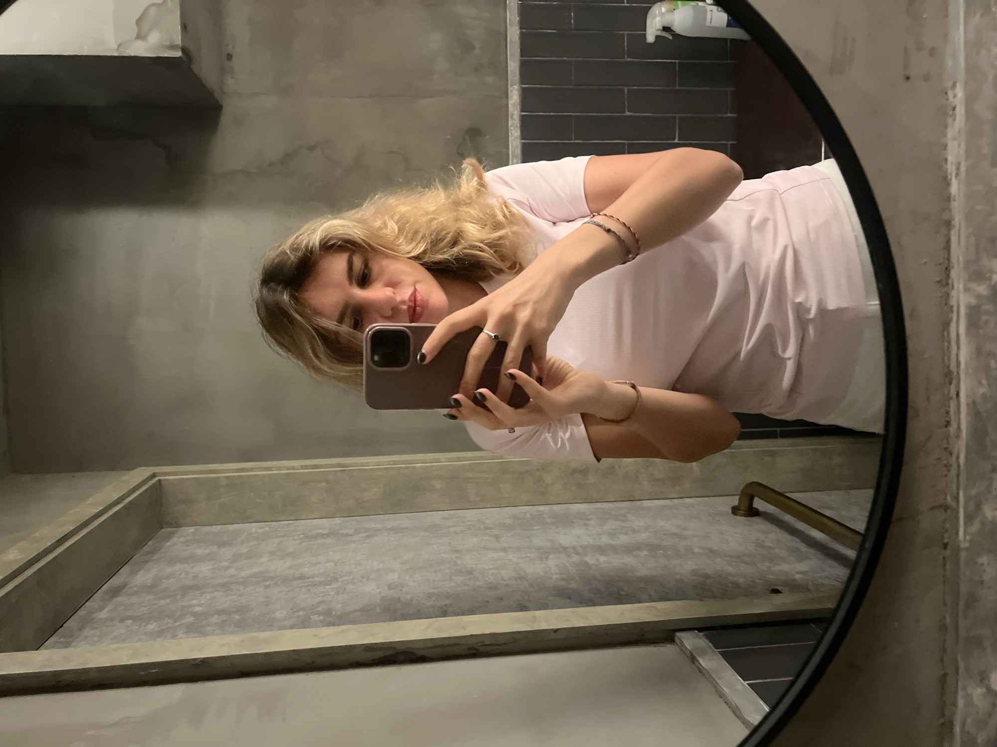The image shows a woman taking a mirror selfie in a bathroom, likely in Hong Kong, China, given the context. She has blonde, wavy hair and is wearing a light pink t-shirt and white shorts. She is holding a smartphone in her right hand and has a ring on her left ring finger. Her nails are painted black. She is wearing two thin bracelets on her left wrist. The bathroom has a minimalist, industrial aesthetic with concrete walls and dark grey tiled backsplash. A gold grab bar is visible in the reflection on the left side of the mirror. On the right side of the mirror, a spray bottle can be seen on a wooden surface. The lighting is dim, creating shadows on the walls.