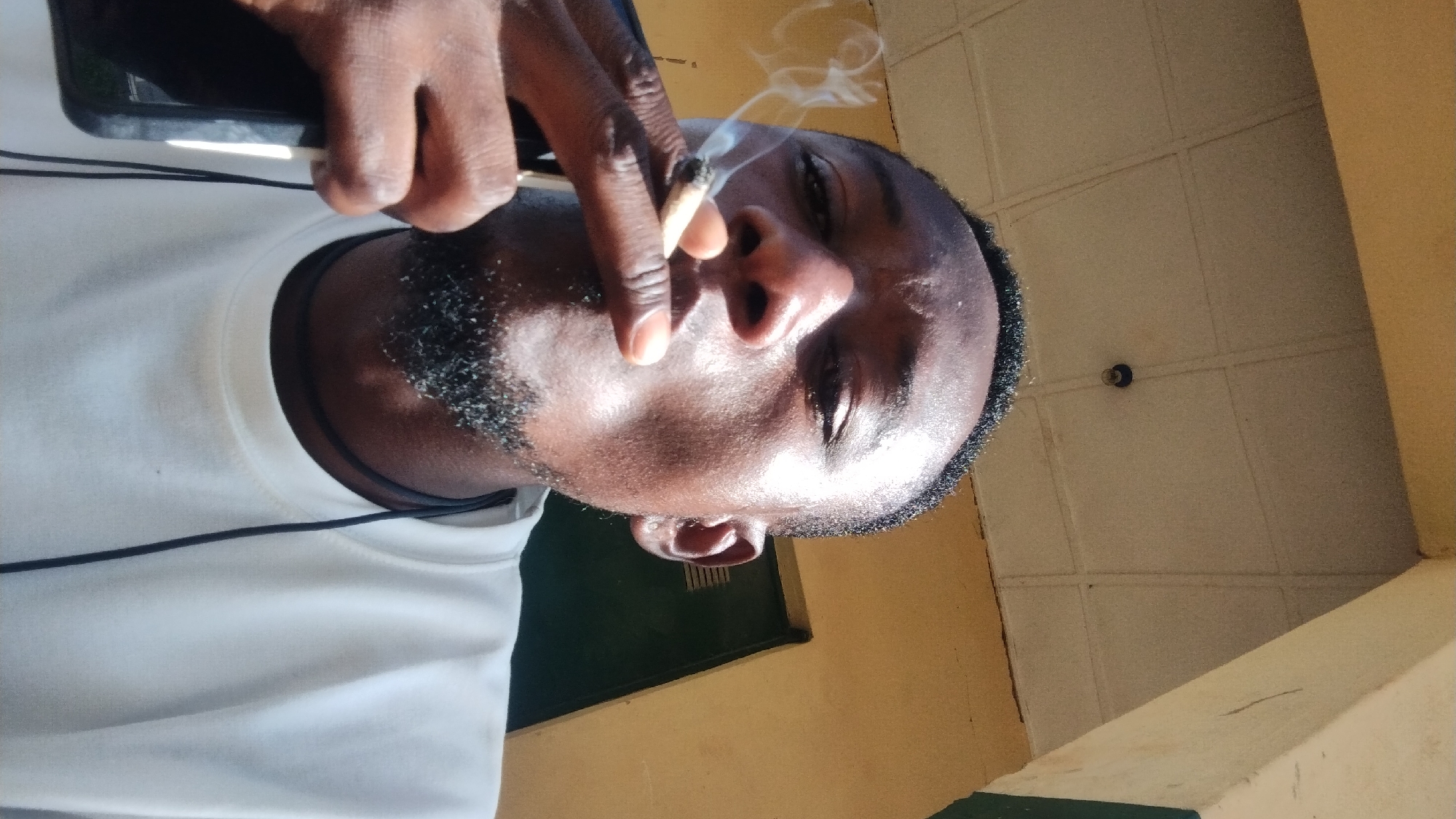 This media file is a close-up, head-and-shoulders shot of a Black man, seen from a low angle. He is holding a phone in his left hand and smoking a cigarette, with smoke rising from it. The man is wearing a white t-shirt. A black cord, possibly from earphones, is visible around his neck and draped over his shirt. His facial expression is neutral, and the lighting suggests it is daytime, possibly sunny, given the strong highlights on his skin.
The background includes a yellow-painted wall and a ceiling with a tiled pattern. A small black circular object, possibly a light fixture or camera, is visible on the ceiling. The overall setting appears to be outdoors or in a partially covered area, with elements of a building visible. The presence of the phone and cigarette suggests a casual moment. The provided location context of Jos, Nigeria, implies a possible West African setting. No text is visible in the image.