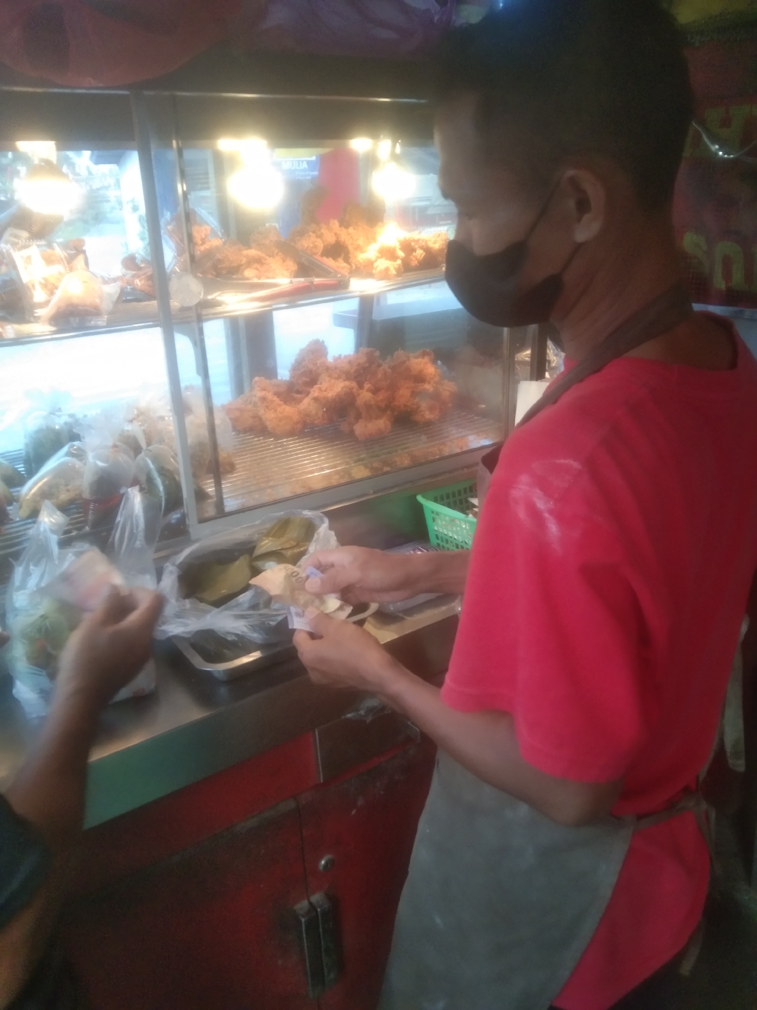 A street food vendor, a man wearing a red shirt and a dark apron, is seen at his stall in Subang, Indonesia, as he handles money exchanged with a customer. The vendor, with dark hair and a black face mask, stands behind a glass display case filled with fried chicken. In the foreground, a customer's hands are visible, holding banknotes and receiving items from the vendor. The stall appears to be an outdoor setup with bright lights illuminating the food and display.