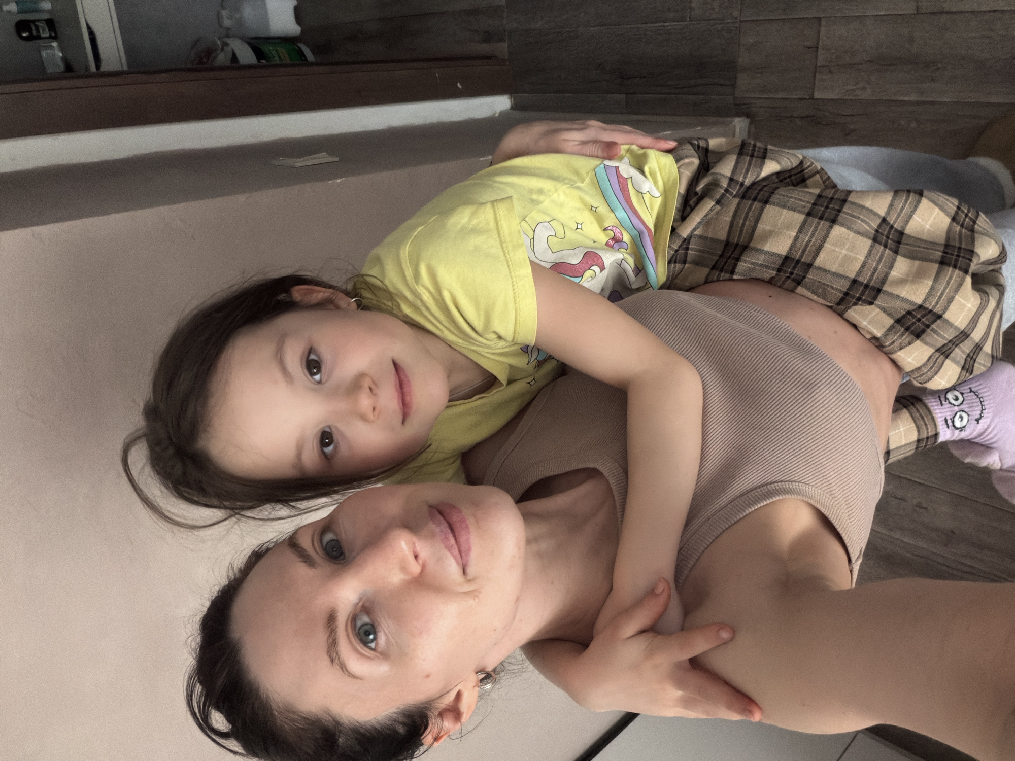 A woman lies on her back while a young girl lies on top of her. The woman has dark hair pulled back, light skin, and blue eyes. She is wearing a beige tank top. The girl has brown hair and brown eyes and is wearing a yellow t-shirt with a unicorn graphic and a brown plaid blanket is covering part of her body. A pale blue blanket is partially visible underneath the plaid blanket. A light-colored wood floor is visible and a light-colored wall. The location is Perm, Russia.
