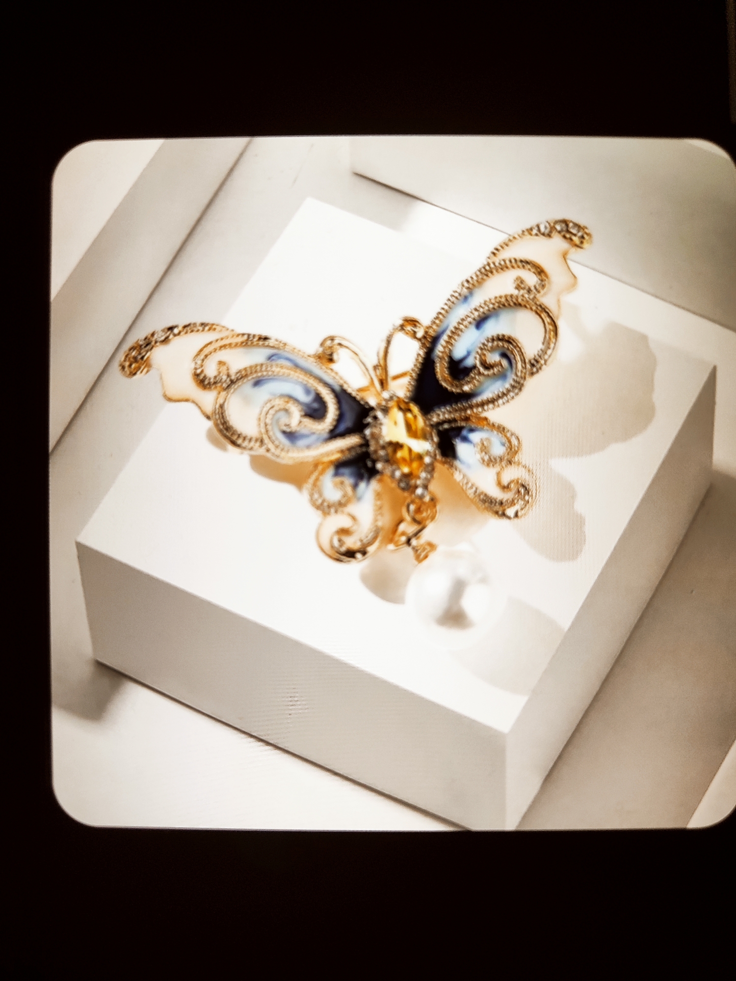 A decorative butterfly brooch with blue and white wings, accented with gold filigree and a yellow gemstone, is displayed on a white cube. A single pearl dangles from the bottom of the brooch. The background is a softly lit display area, with shadows cast by the object. The image has a vintage, slightly blurry aesthetic due to a rounded-corner filter applied. The location is Unknown, Unknown.