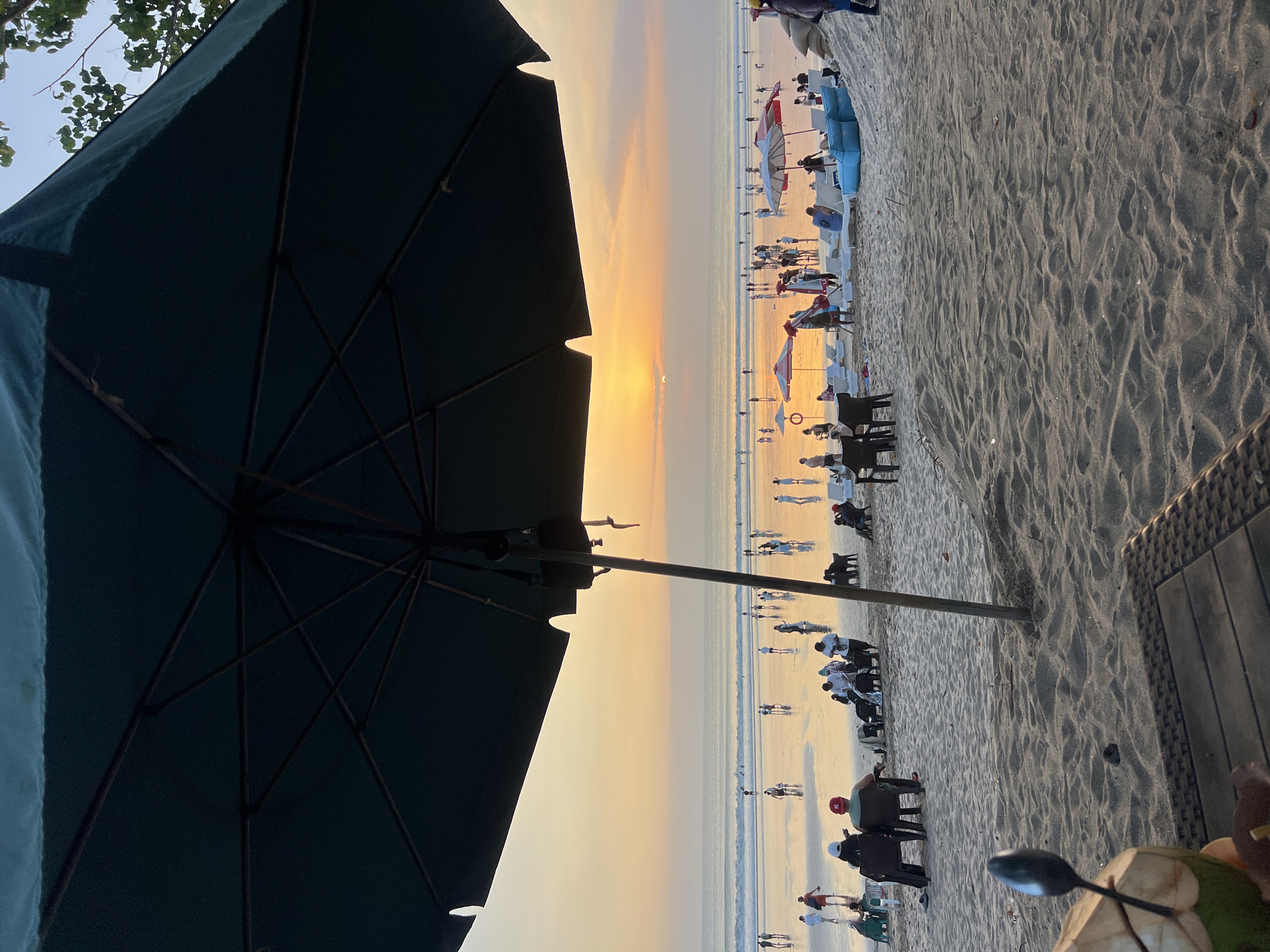 The image depicts a sunset scene on Kuta Beach in Indonesia. In the foreground, a part of a dark green beach umbrella and a table with a coconut and spoon are visible. The midground shows a sandy beach populated with numerous people. Some individuals are sitting on chairs and loungers, while others are walking along the shore or in the shallow water. Several colorful beach umbrellas are set up, and lounge chairs are scattered across the sand. The background features the ocean with gentle waves and the sun setting, casting a warm orange glow across the sky and reflecting on the wet sand. The overall atmosphere is one of a relaxed beach environment during twilight.