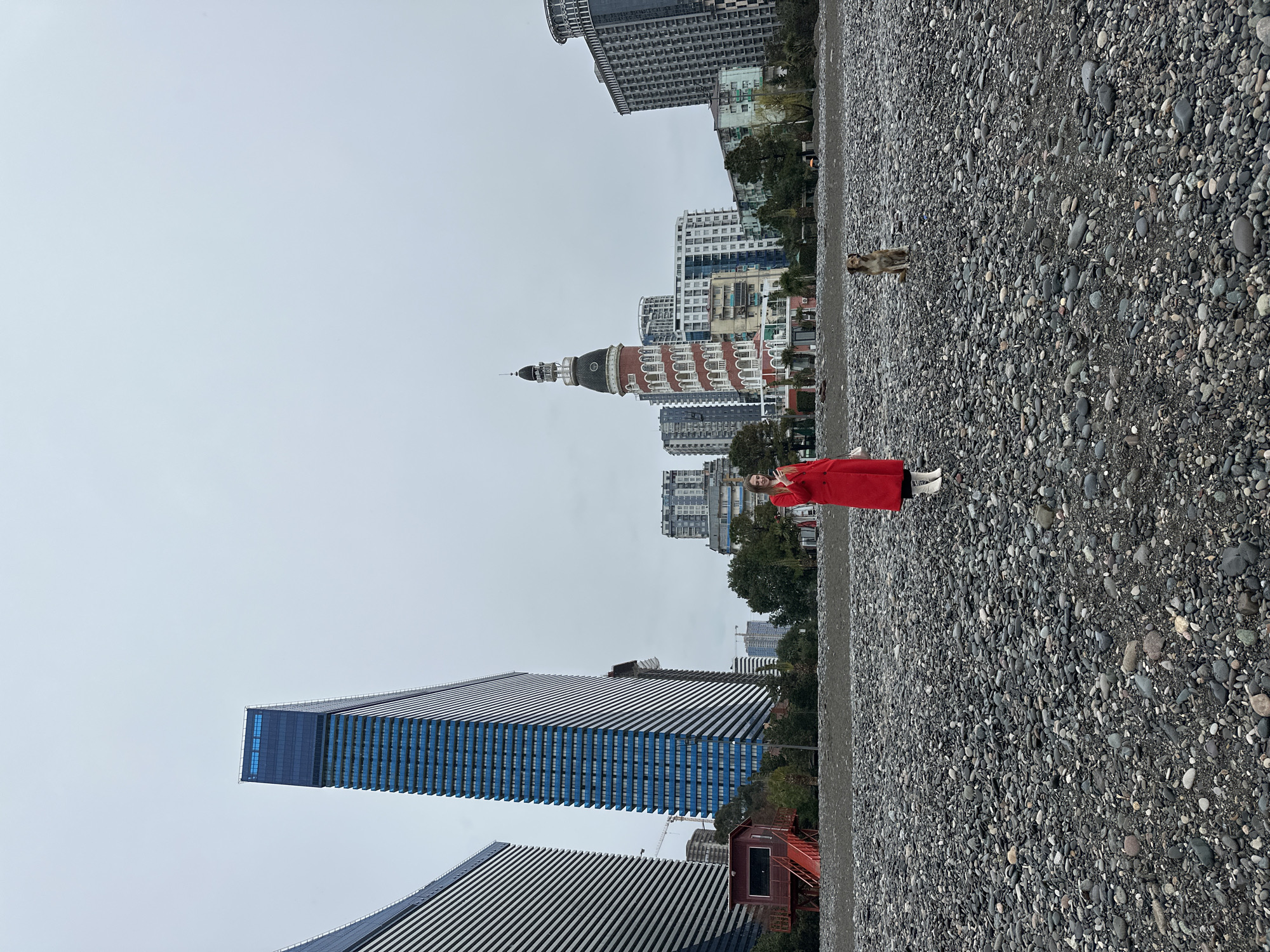 The photograph depicts a person wearing a bright red coat and white boots standing on a pebble beach in Batumi, Georgia. In the background is a skyline of modern and older architectural buildings, including the iconic Alphabet Tower. A dog is positioned in the middle of the beach, further back. The sky is overcast. The beach consists entirely of gray, smooth stones of varying sizes.