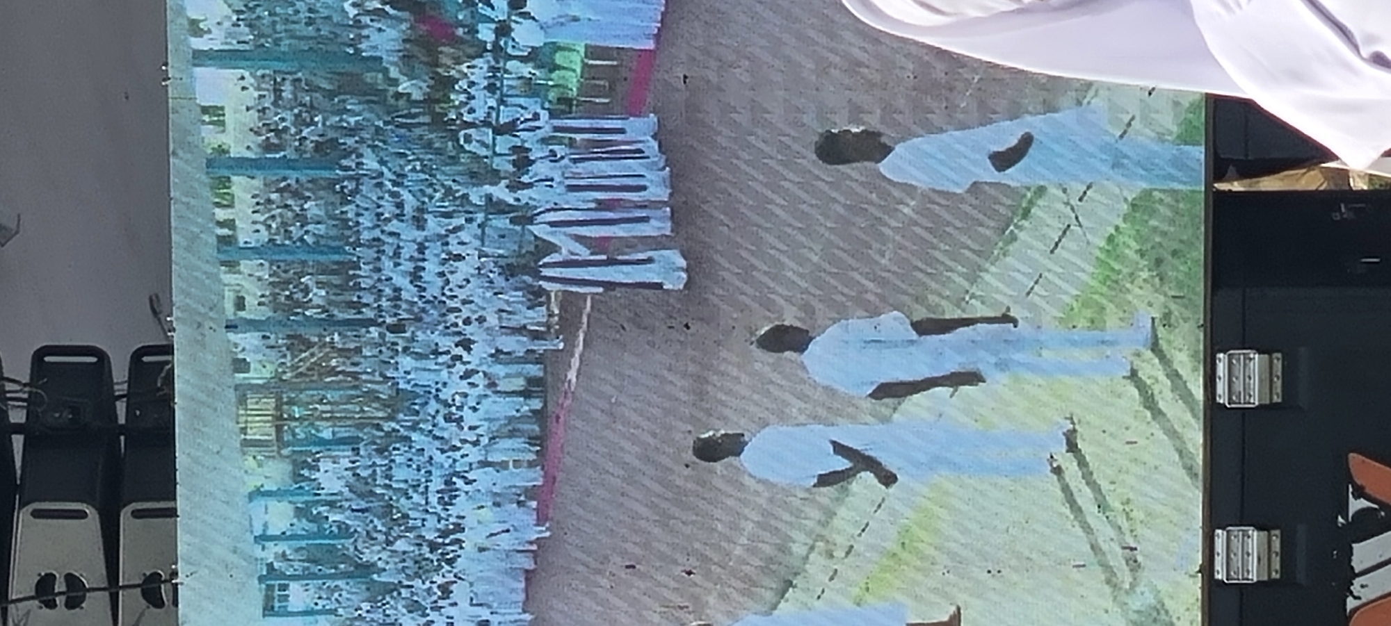 The image shows a monitor displaying a large group of people in white uniforms, seemingly on a parade ground. In the foreground, there are individuals also in white uniforms marching in formation. The background appears to show a building and a large crowd. The setting is suggested to be Calabar, Nigeria, based on the prompt. Additional chairs and equipment are visible at the edge of the frame on the left side, and hardware for mounting or attaching the monitor is seen on the right.