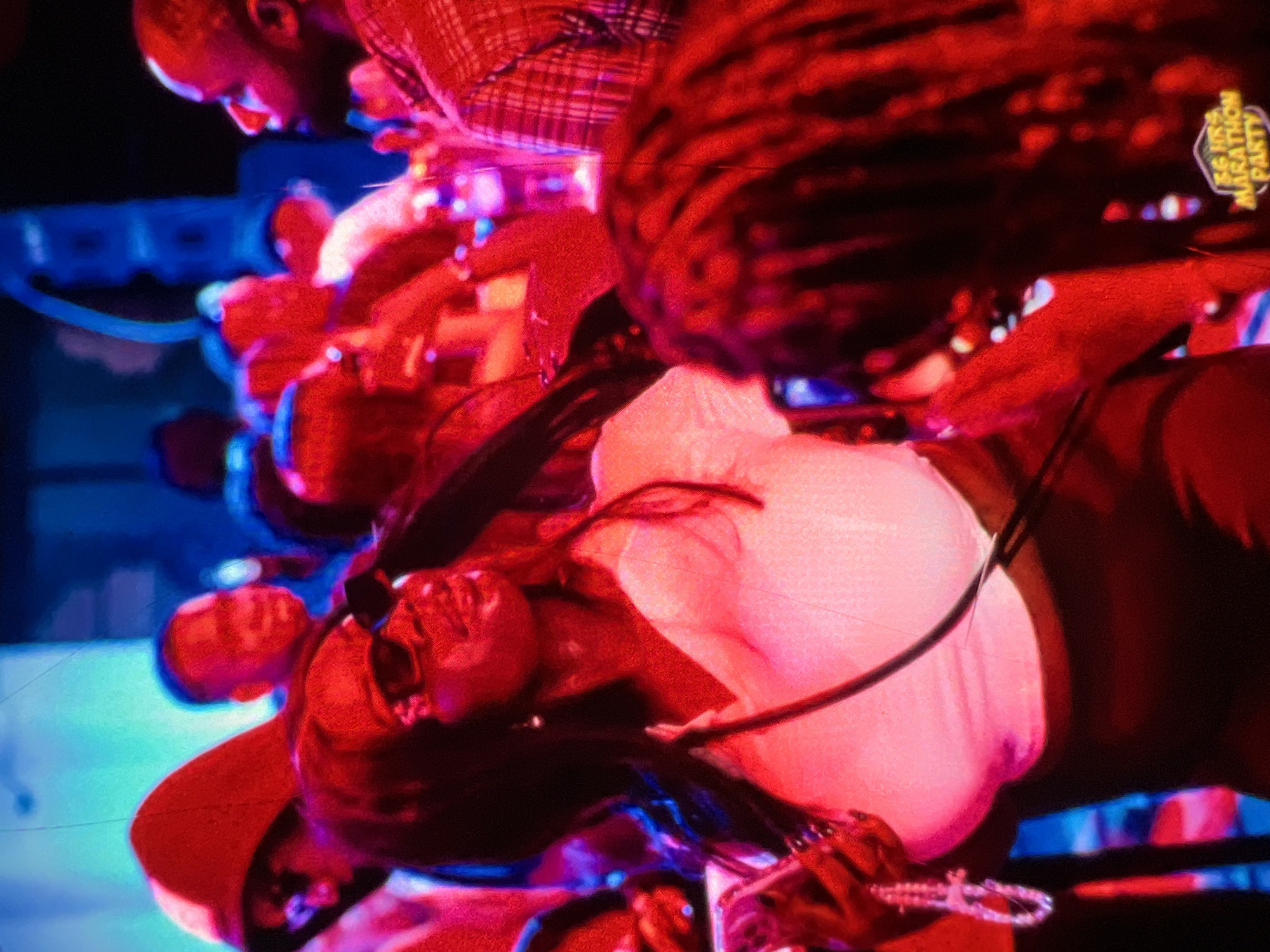 The image depicts a crowded indoor scene, likely a party or event, with participants in what appears to be Lagos, Nigeria, suggested by the context of the marathon party sticker visible on the right side of the image. The lighting is predominantly red and blue, creating a vibrant and somewhat dramatic atmosphere.
In the foreground, a woman with long dark hair and sunglasses is the central focus. She is wearing a white top with a deep V-neckline and a dark skirt. She is holding a smartphone in her left hand and has a beaded bracelet on her wrist. To her right, another person's head and shoulders are visible, featuring dreadlocks and a sticker that reads "56 HRS MARATHON PARTY".
Behind the main subject, other individuals are partially visible. To her left, a person in a red hooded jacket and sunglasses is looking to the side. Further back, other blurry figures are present, some with illuminated faces from the colored lighting. The background is a mix of dimly lit areas and colored light projections, suggesting a performance space or a decorated venue. The overall impression is of a lively social gathering.