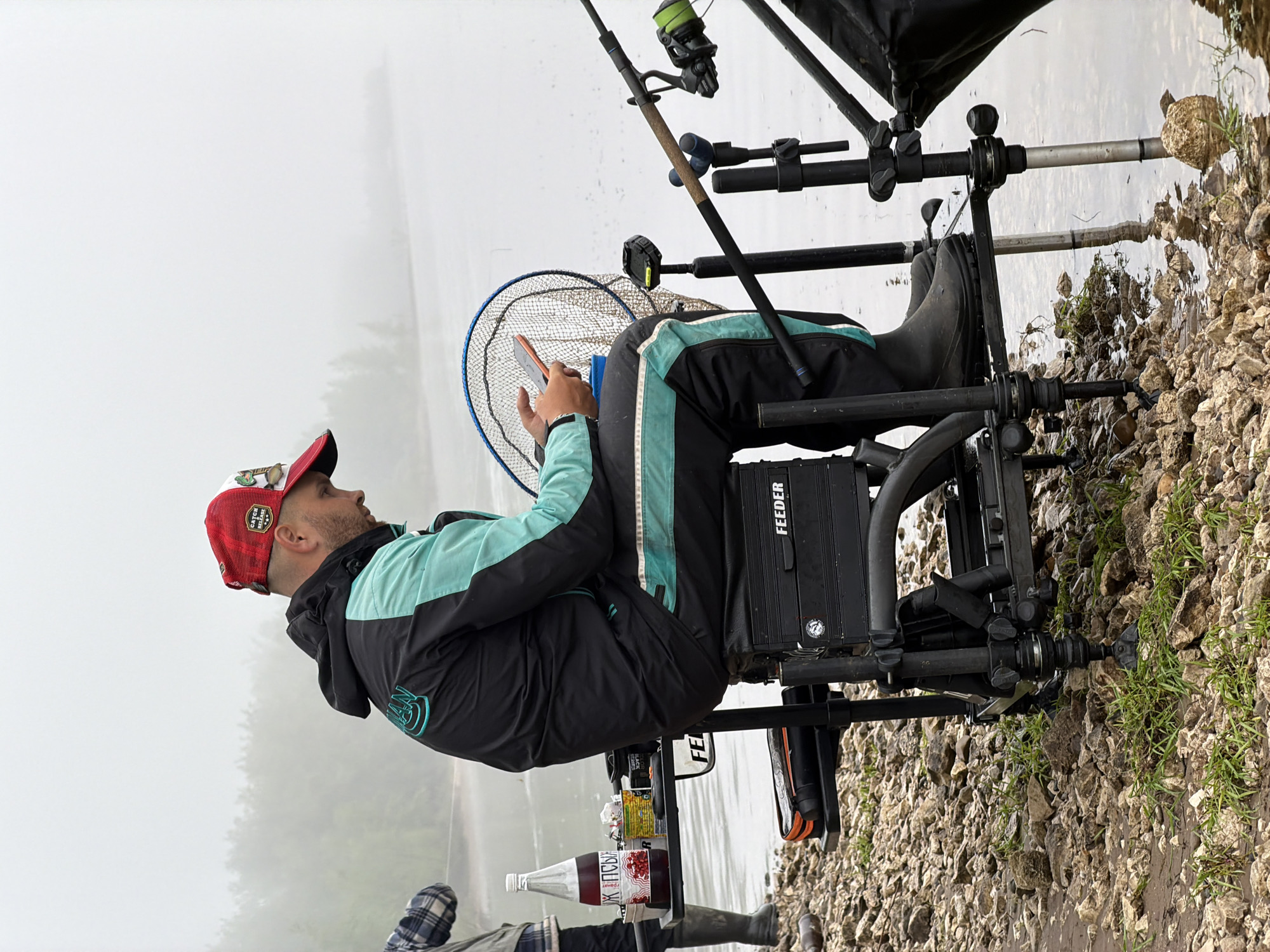 This image, taken in Chaikovsky, Russia, captures a man engaged in feeder fishing by a body of water on a very foggy day. The man is positioned on the right side of the frame, seated on a specialized fishing chair or platform, facing left towards the obscured water. He wears a red and white baseball cap with "CATCH & RELEASE" visible on its front, a black and teal jacket, and dark pants.
In his hands, he holds a small orange device, possibly a phone, and appears focused on it or the water ahead. Surrounding him is a comprehensive fishing setup: multiple fishing rods with reels are mounted on a rod pod or stand directly in front of his seat. A large fishing net rests on his lap. A black box or bag, prominently labeled "FEEDER," is attached to his chair. Nearby, a bottle containing a red liquid, with Cyrillic text "Я РІРБІСУ" (likely "Cranberry"), is also attached to his setup.
The setting is a rocky and gravelly shoreline, with sparse green grass growing among the stones. The water's surface reflects the soft, diffused light of the sky. The most notable detail is the pervasive, dense fog or mist that heavily obscures the background, making distant trees and the opposing shore nearly invisible. The diffused lighting suggests an early morning, late afternoon, or simply a heavily overcast day, contributing to a tranquil and contemplative atmosphere.