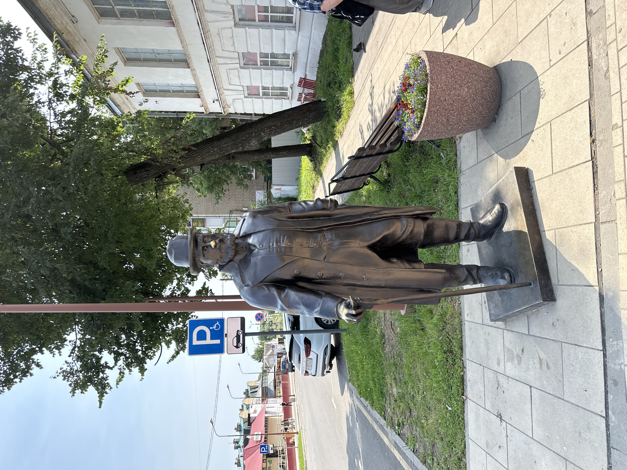 A bronze statue of a man in a top hat and long coat stands on a paved sidewalk in front of a white building with windows and a red-tiled roof in Selivanovo, Russia. The man holds a cane and is positioned to the left of a large, circular planter filled with flowers. A wooden bench is visible behind the planter, and a tree with green leaves grows behind the bench. A street with a white car and a parking sign is to the left of the statue. The sky is blue with scattered clouds.