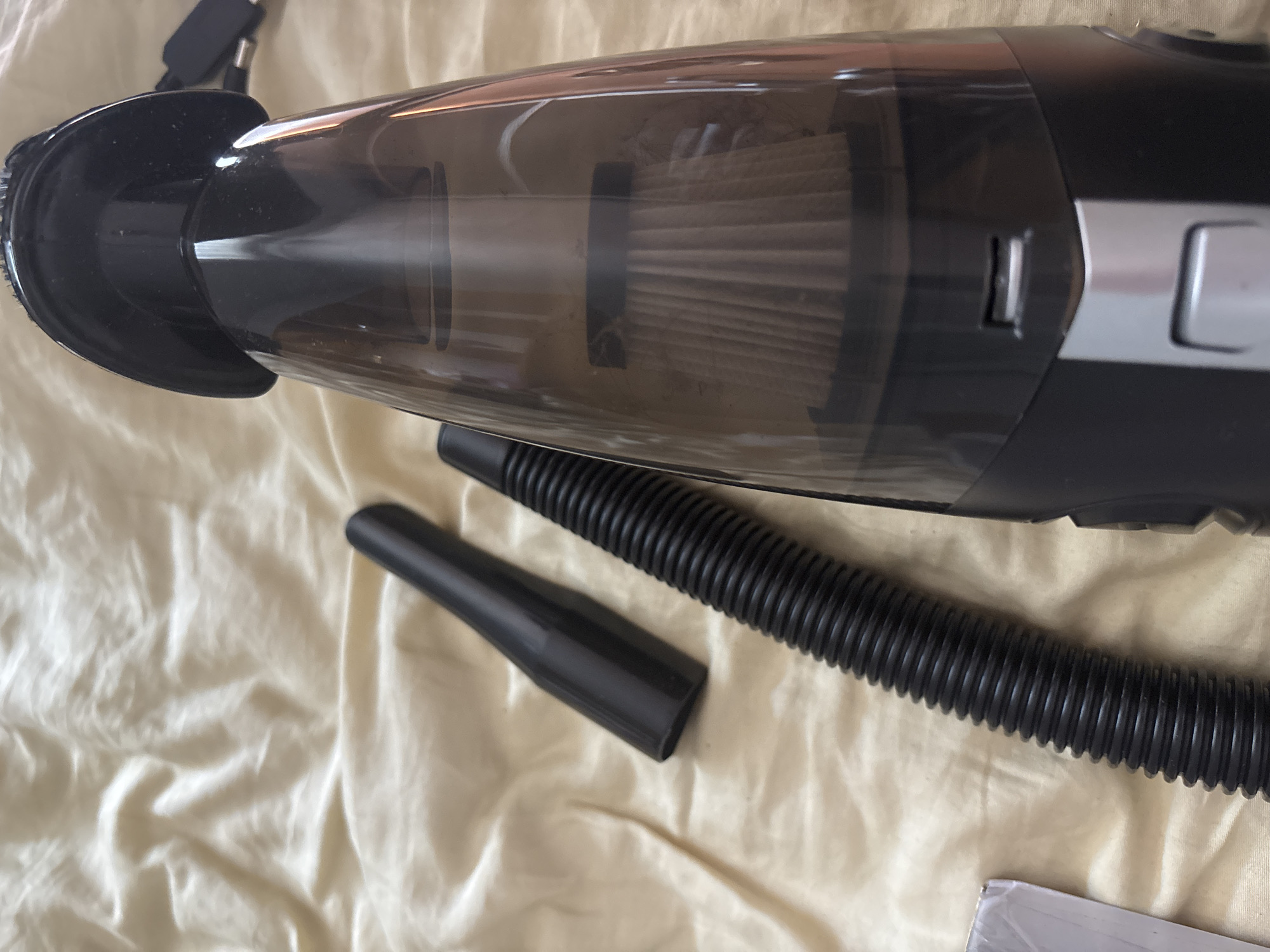 The image shows a close-up, overhead view of a handheld vacuum cleaner and its accessories, laid out on a creased, light-colored fabric, possibly a bedsheet. The vacuum cleaner is black with a transparent dustbin, revealing a pleated filter inside. A black, ribbed hose and a black, cone-shaped nozzle are positioned below the vacuum. The scene is lit with artificial light, casting soft shadows and highlighting the texture of the fabric. There is no visible text in the image.