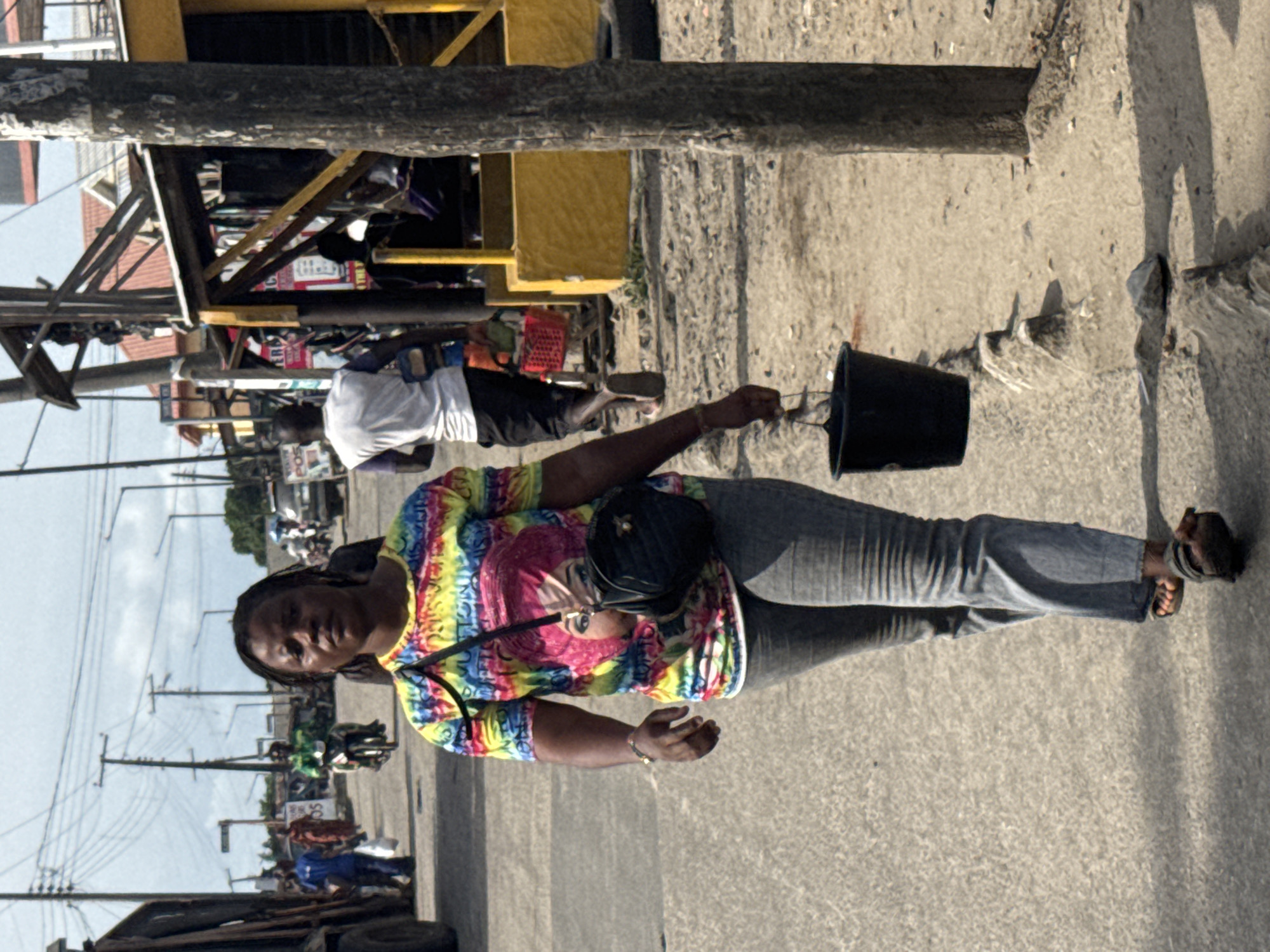 A woman walks on a city street in Badagry, Nigeria, carrying a black bucket. She wears a multicolored t-shirt, blue jeans, and sandals. A black purse hangs from her shoulder. There are other people in the background and a yellow structure above. Powerlines are visible in the distance.