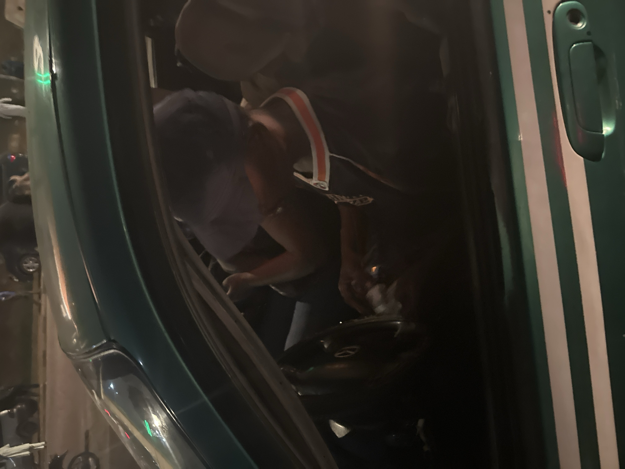 The image shows the interior of a dark green car with white stripes, likely a taxi. A person wearing a blue baseball cap and a black shirt with an orange and white striped collar is seated in the driver's seat, looking down at something in their hands. The steering wheel is visible, and the car appears to be stopped, possibly in traffic. The background shows a blurred nighttime street scene with other vehicles and pedestrians, suggesting an urban environment like Abuja, Nigeria. The lighting is dim, and the overall atmosphere is that of a busy city at night.