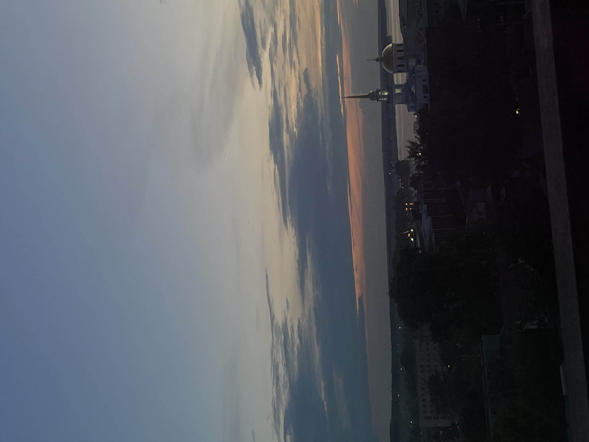 The image depicts a sunset over a body of water, likely a river or lake, in Votkinsk, Russia. Silhouetted against the colorful sky are buildings, including a prominent structure with a golden dome and a tall, slender spire, characteristic of a church or cathedral. The sky transitions from a pale blue at the top to hues of orange and pink near the horizon, indicating the end of the day. Wispy clouds are scattered across the sky, adding texture and depth to the scene. In the foreground and on the right side of the image, the view is partially obscured by dark shapes, possibly trees or a balcony railing. The overall mood is tranquil and serene, with the fading light casting long shadows and bathing the landscape in a warm glow. There is no visible text in the image.
