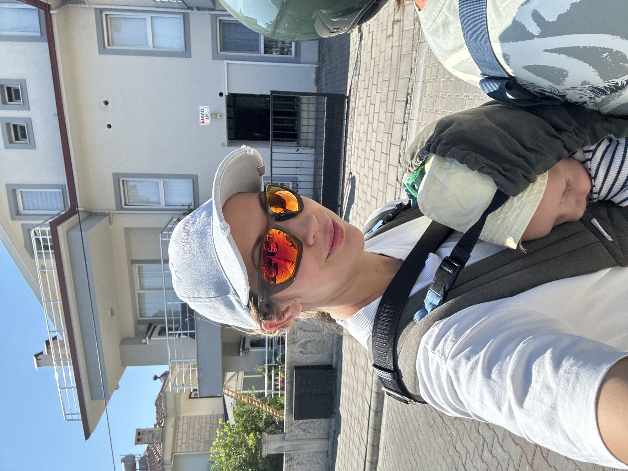 A woman wearing a white t-shirt, a light blue baseball cap, and sunglasses with orange lenses is taking a selfie while riding a scooter. She is wearing a dark grey baby carrier on her front, and a baby is asleep inside, wearing a light beige hat and a striped shirt. The woman is looking at the camera with a slight smile. In the background, there is a modern, light grey building with white window frames and balconies. A sign on the building reads "KARAYEL APT." The street is paved with brick-like stones. The scene suggests a sunny day in a residential area of Fethiye, Turkey, possibly near a coastal town due to the clear blue sky.