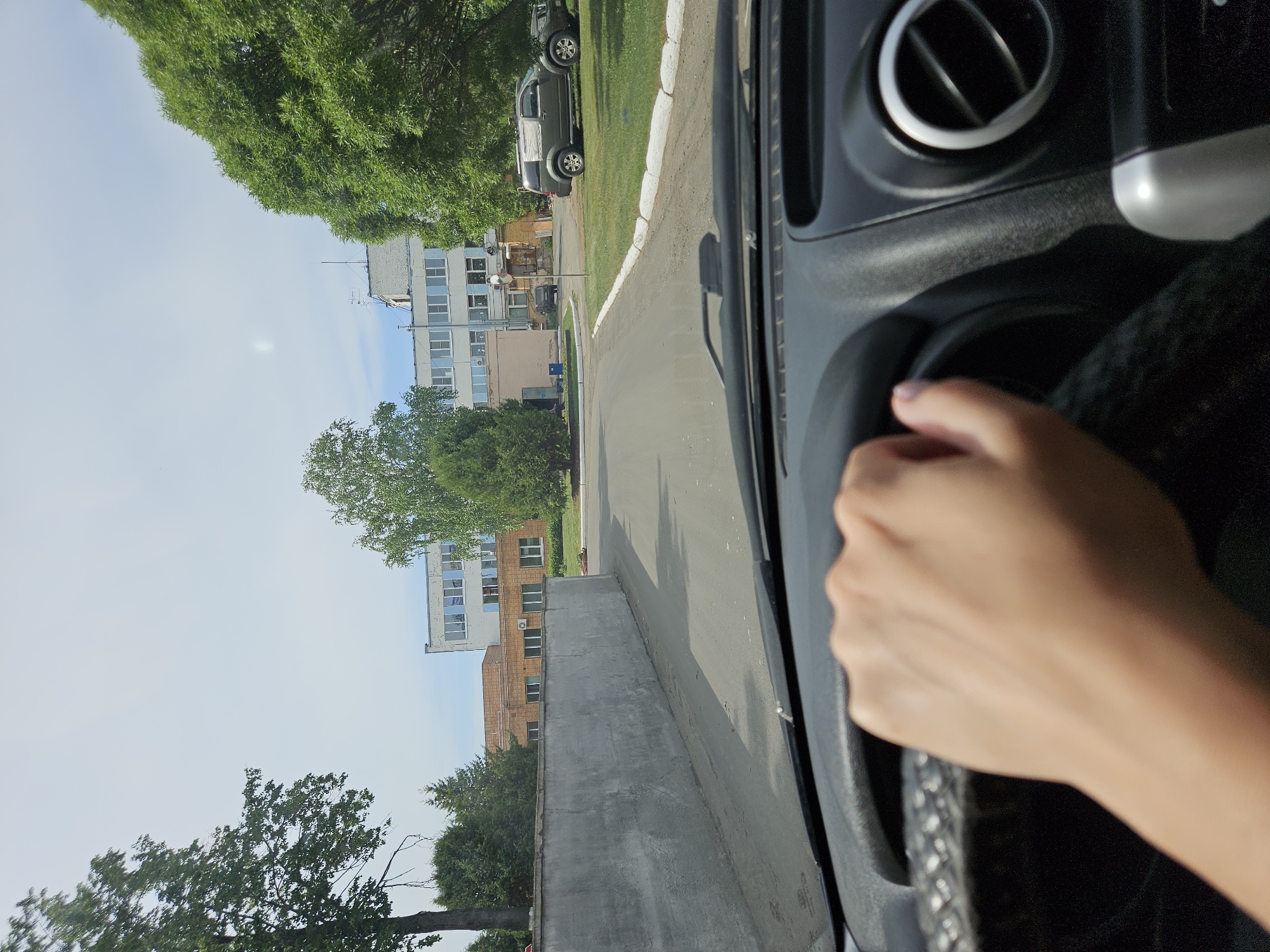 A person is driving a car on a paved road in Chaikovsky, Russia. The driver's left hand is visible on the steering wheel. On the left side of the road is a low concrete wall, and beyond that, there are multi-story buildings and trees. Another car is parked on the right side of the road. The sky is visible above the buildings and trees. The car's dashboard and air vent are visible on the right side of the frame.