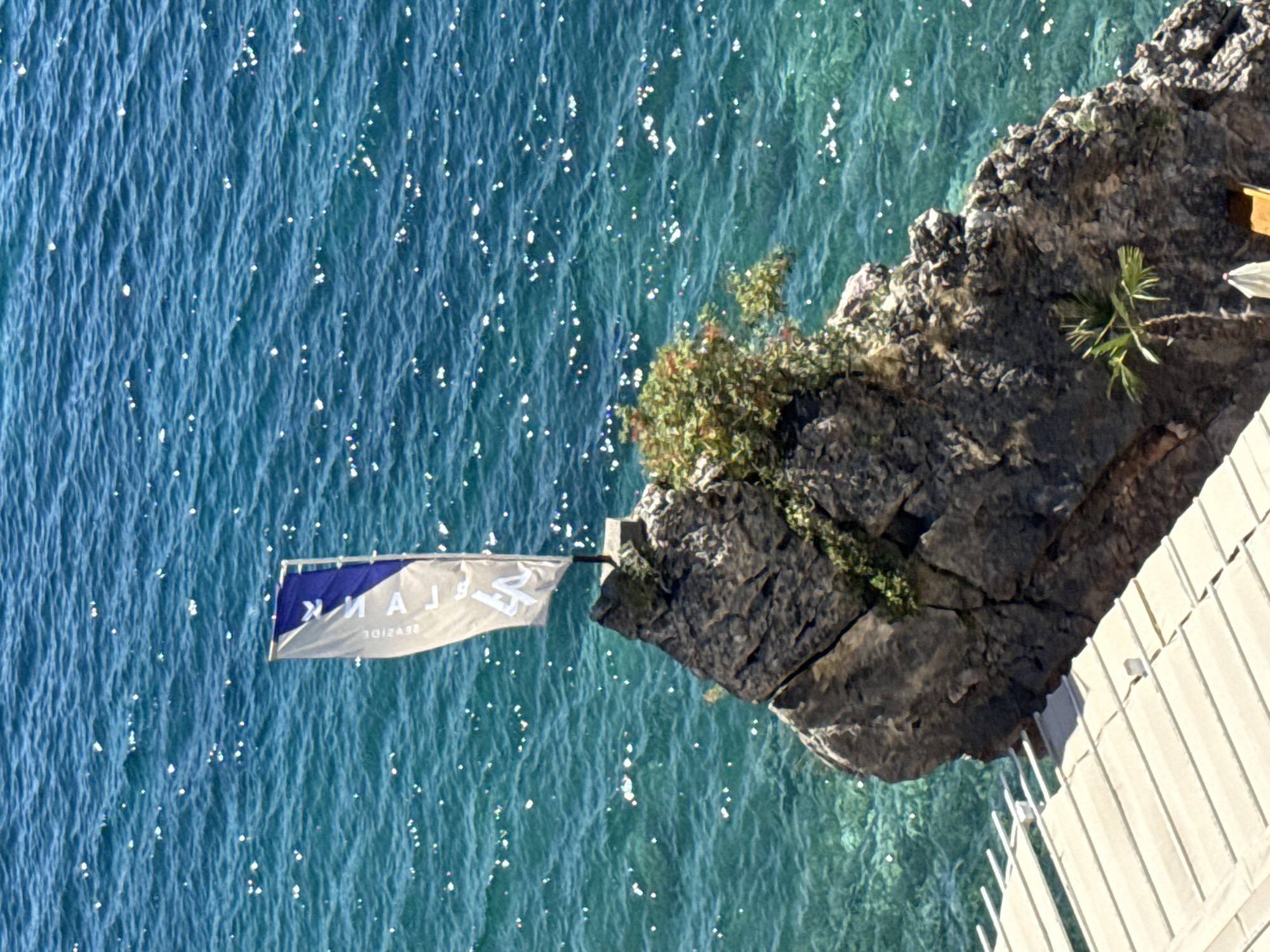 A flag with a blue and white pattern flies from a rocky outcrop in the turquoise waters of Budva, Montenegro. The flag has text in a mirrored fashion, reading "2EVJIDE" and "MAJ", suggesting it is a personal or business flag. The rocky outcrop is sparsely vegetated with a small palm tree at its base. A portion of a wooden structure, possibly a pier or deck, is visible in the foreground. The water is clear and sparkling, with sunlight reflecting off its surface.